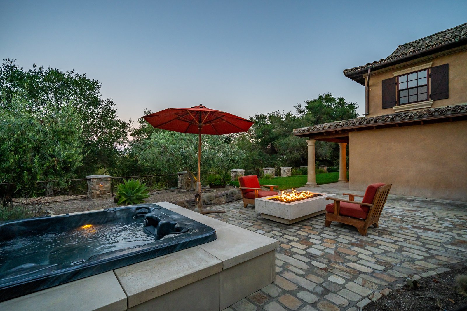 Outdoor patio with a hot tub, fire pit with seating, red umbrella, and lush greenery in the background.
