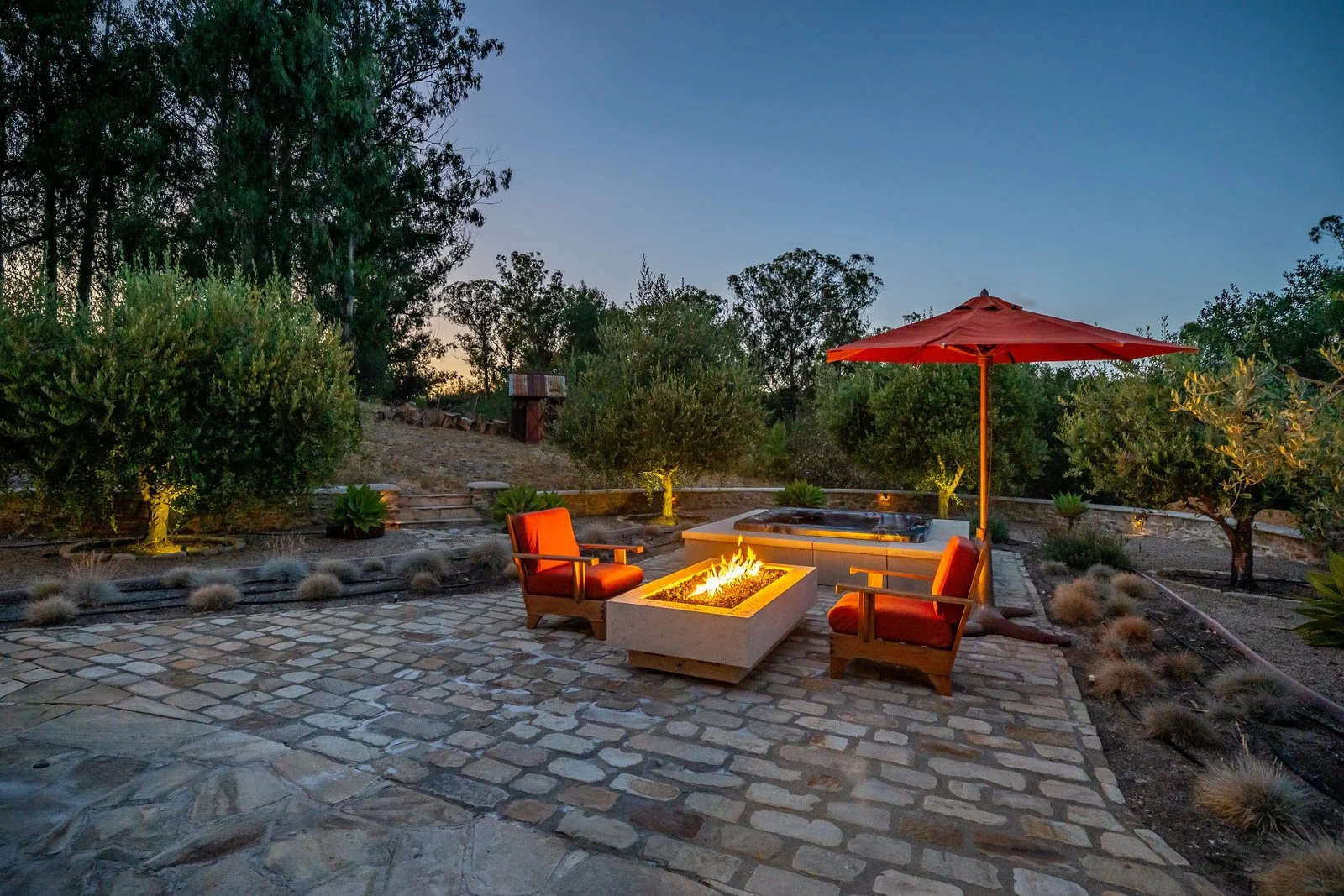 Outdoor patio scene at dusk with two orange cushioned chairs around a rectangular fire feature, a hot tub with a red umbrella, surrounding trees, plants, and a cobblestone patio.