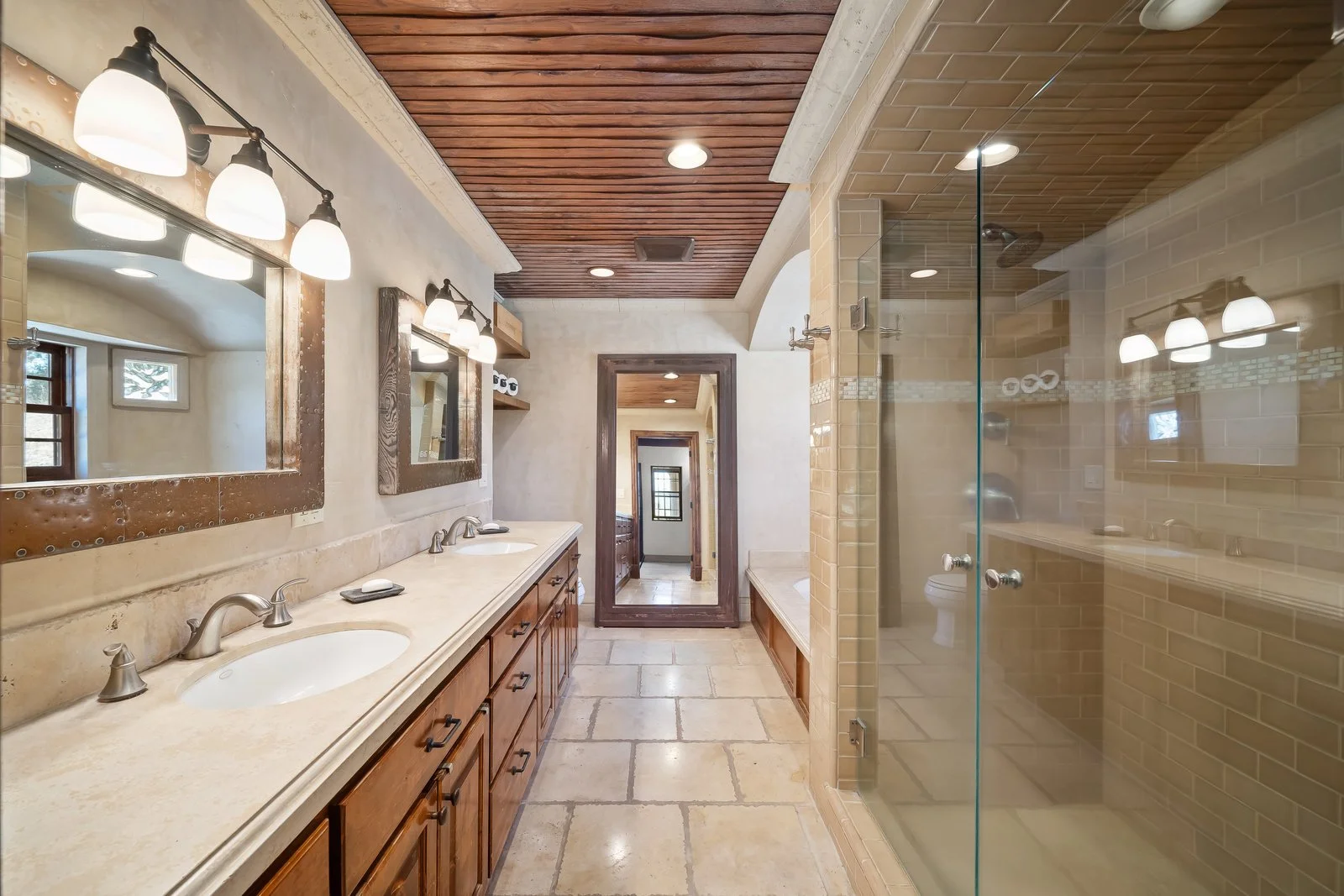 A spacious bathroom with dual sinks, wooden cabinets, and large mirrors. There are two wall-mounted light fixtures above the mirrors. The bathroom has beige tiled floors, a glass shower with yellowish tiles, and a wooden ceiling. A doorway leads to a