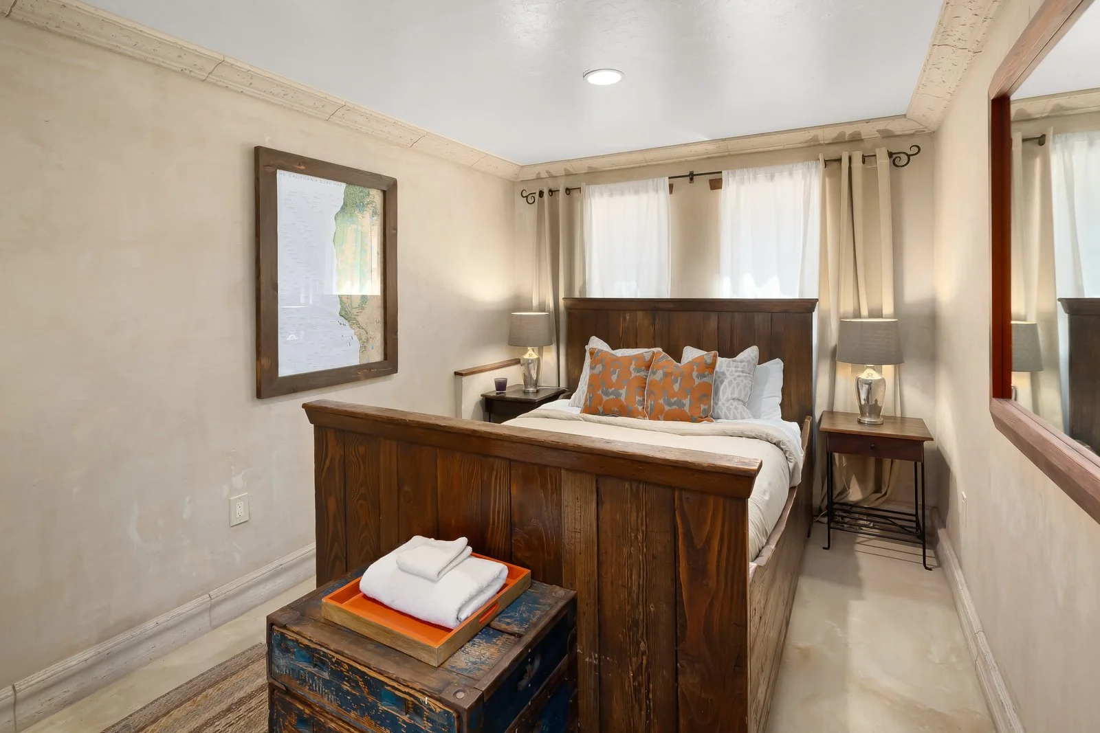 A cozy bedroom with a wooden bed frame, white bedding, and decorative pillows. Two nightstands with lamps, a framed map on the wall, and cream curtains on windows.