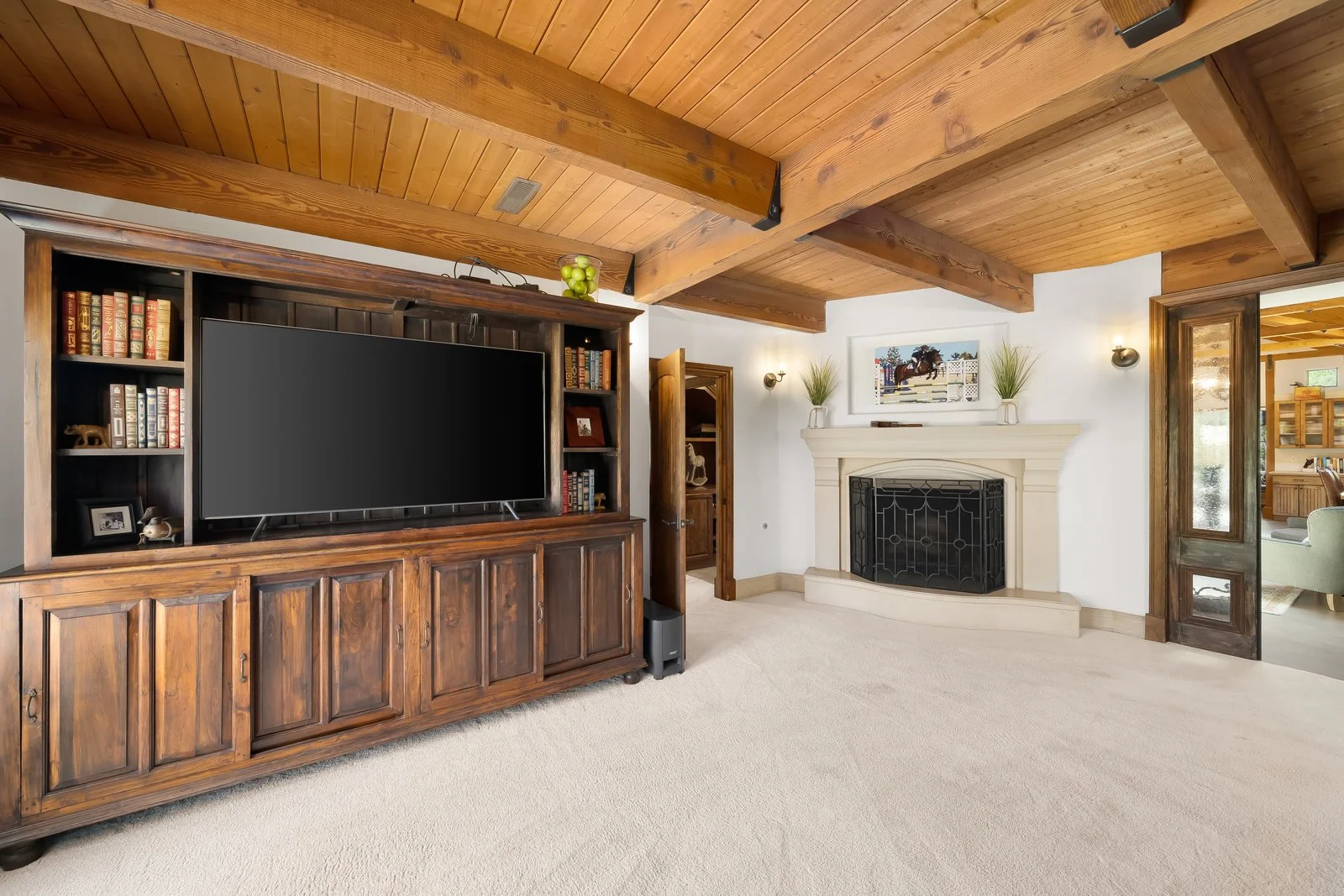 Living room with wood ceiling, a large wooden entertainment center with a flat screen TV, fireplace with mantel, wall sconces, and decorative plants on the mantel, open doorway revealing a sitting area.