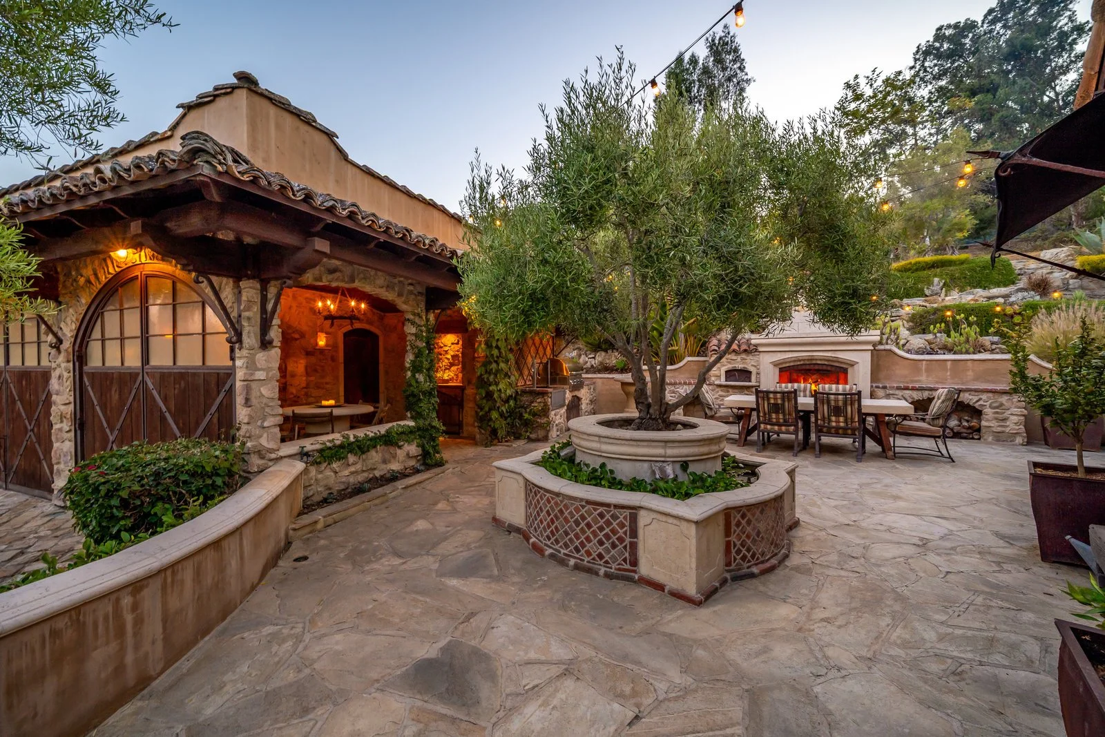 An outdoor patio area with a stone floor, surrounded by greenery and trees. There is a central large round planter with a tree, a fireplace built into a stone wall, outdoor dining table with chairs, a small covered seating area, and string lights han
