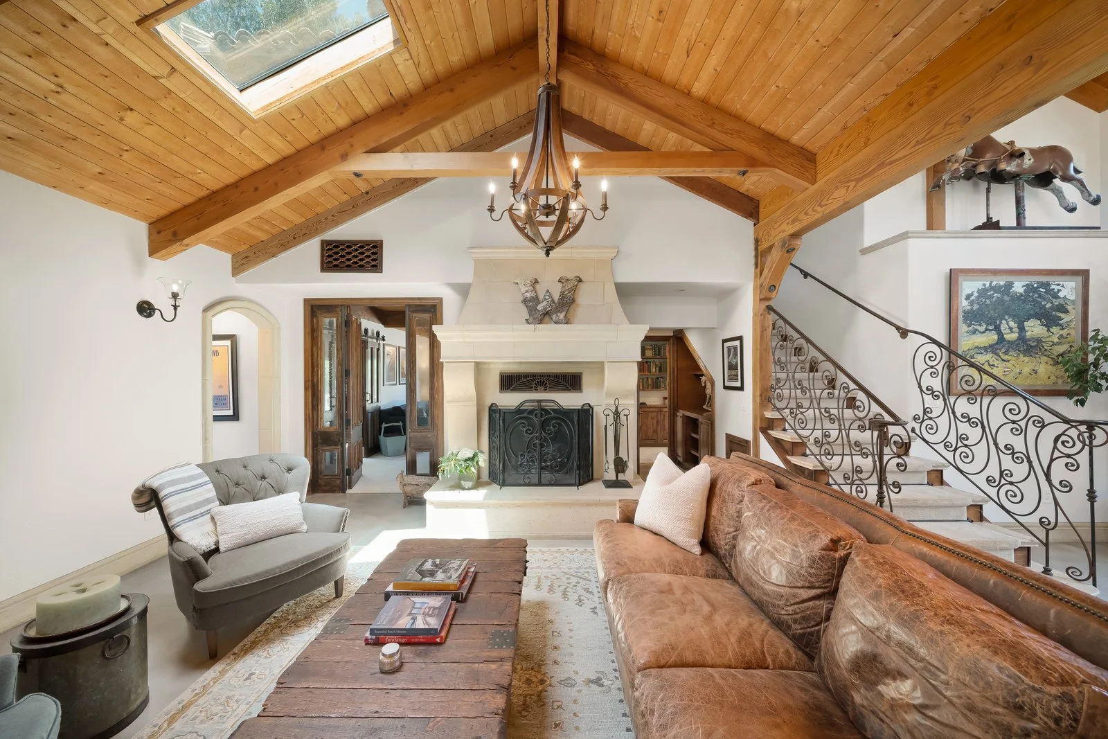 Living room with a wooden vaulted ceiling, a skylight, fireplace, and antique furniture including a leather sofa and a tufted armchair. Decor includes artwork, a chandelier, and a staircase with ornate iron railing.