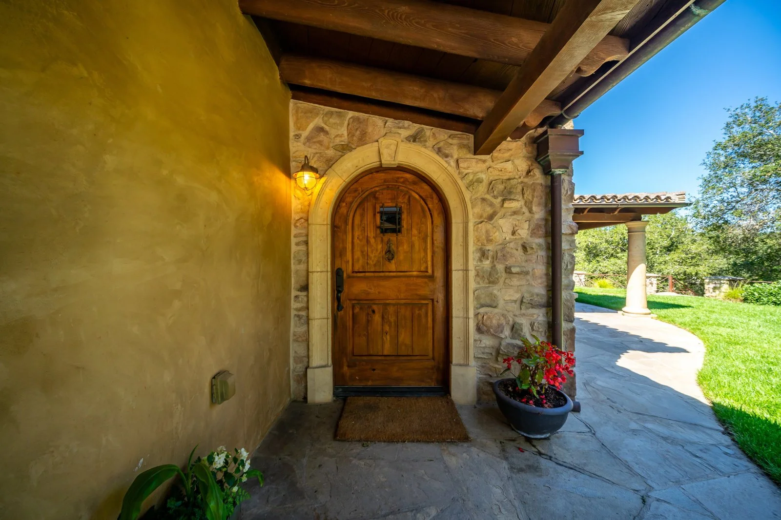 Wooden front door with small window, surrounded by stone arch and walls, outside porch with potted flowers and grassy lawn on a sunny day.