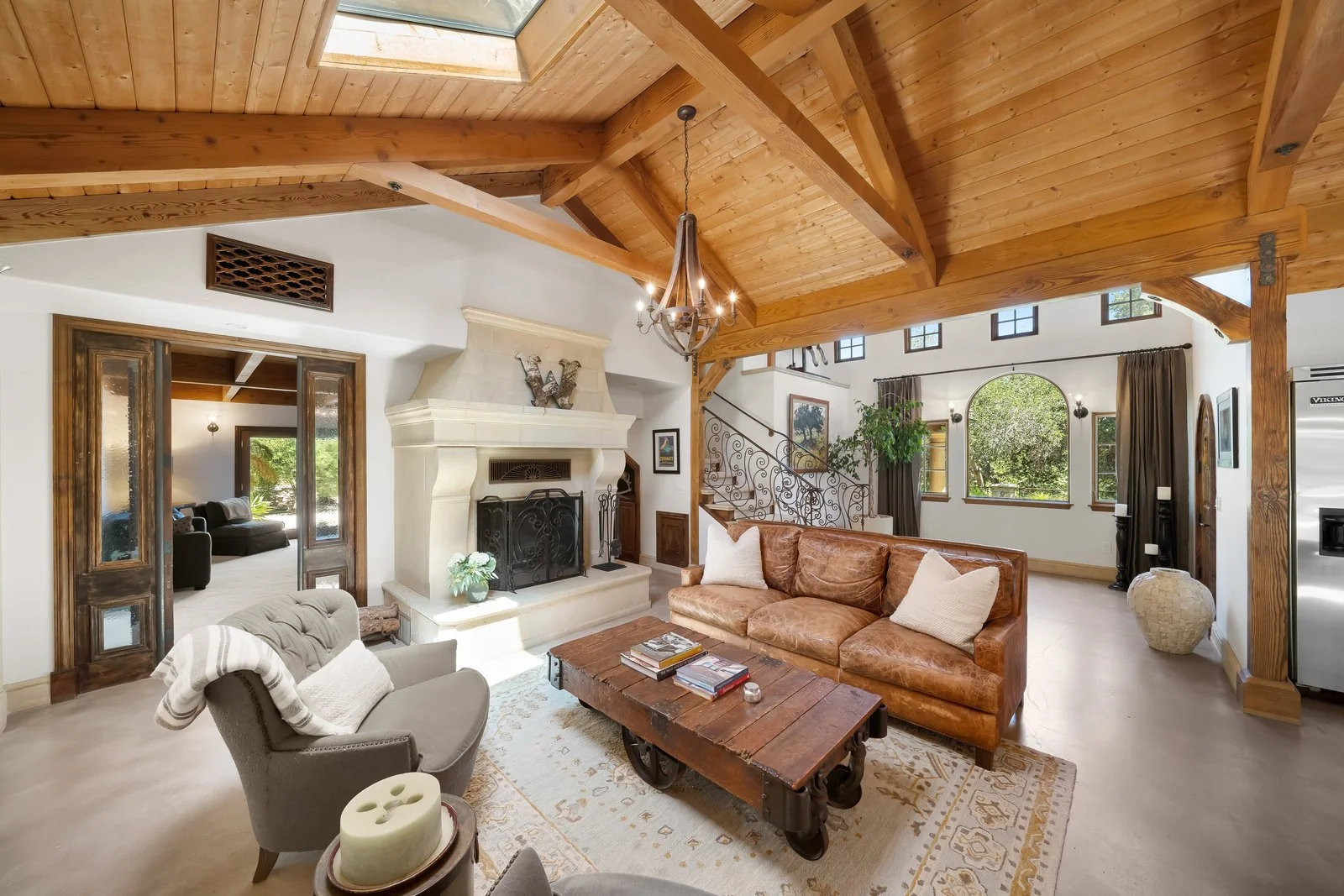 Living room with wooden vaulted ceiling, stone fireplace, brown leather sofa, gray armchair, rustic wooden coffee table, and large arched window with curtains.