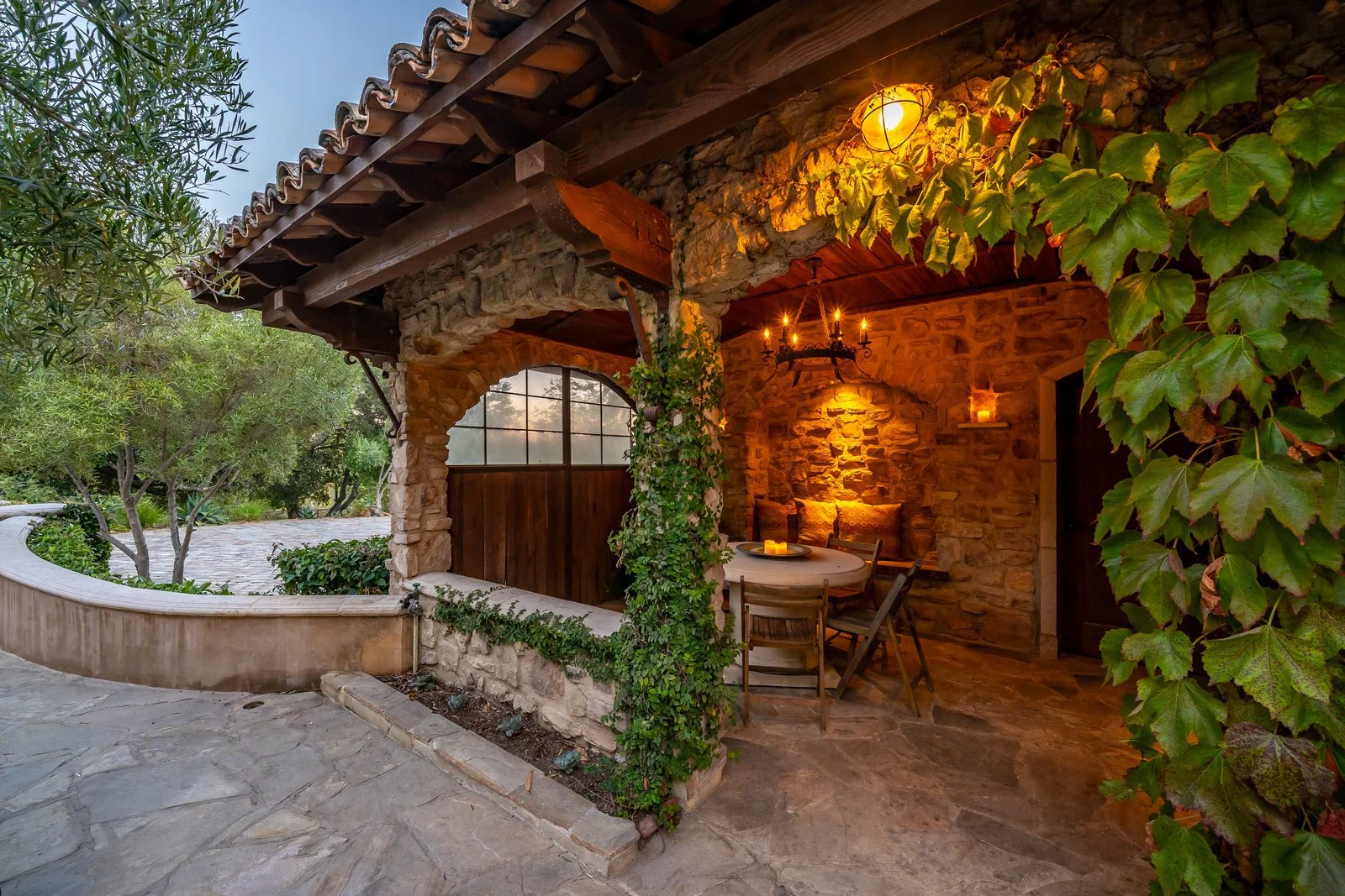 Cozy outdoor patio with stone walls and wooden furniture, illuminated by warm lighting and candles, surrounded by greenery.