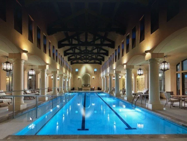 Indoor swimming pool with lane markings, surrounded by lounge chairs and ornate lantern-style lighting, in a luxurious setting with high vaulted ceilings and columns.