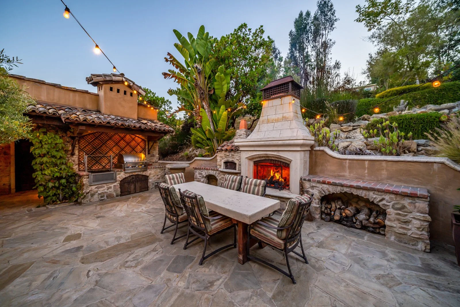 Outdoor patio with stone flooring, a large fireplace with a fire burning, a long table with six chairs, string lights overhead, green plants, and a barbecue grill in a backyard setting at dusk.