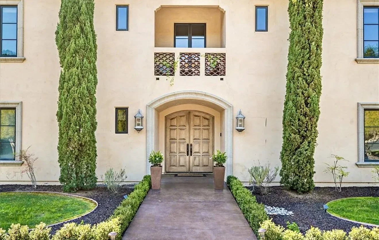 Front view of a beige residential building with a double wooden door, flanked by two tall green trees, and decorated with outdoor lantern lights, potted plants, and a landscaped path and garden.