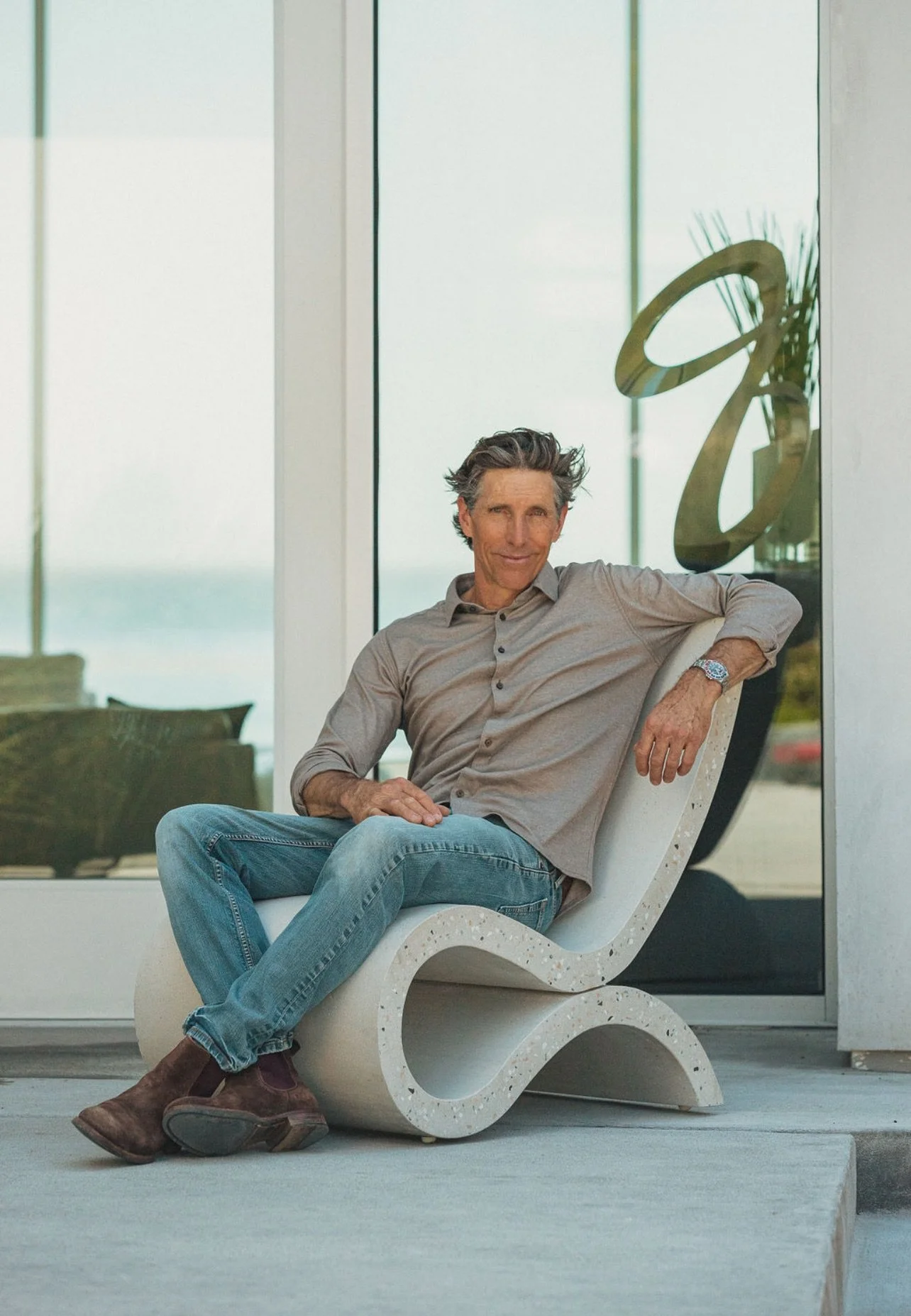 Steffen Weeks sitting comfortably in a modern, sculptural chair he hand crafted outside large glass doors, with a gold decorative element in the background.