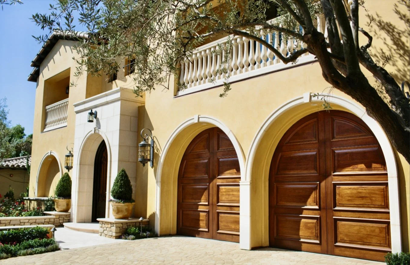 Exterior of a yellow Mediterranean-style house with two wooden garage doors, arched entryway, potted plants, and trees.