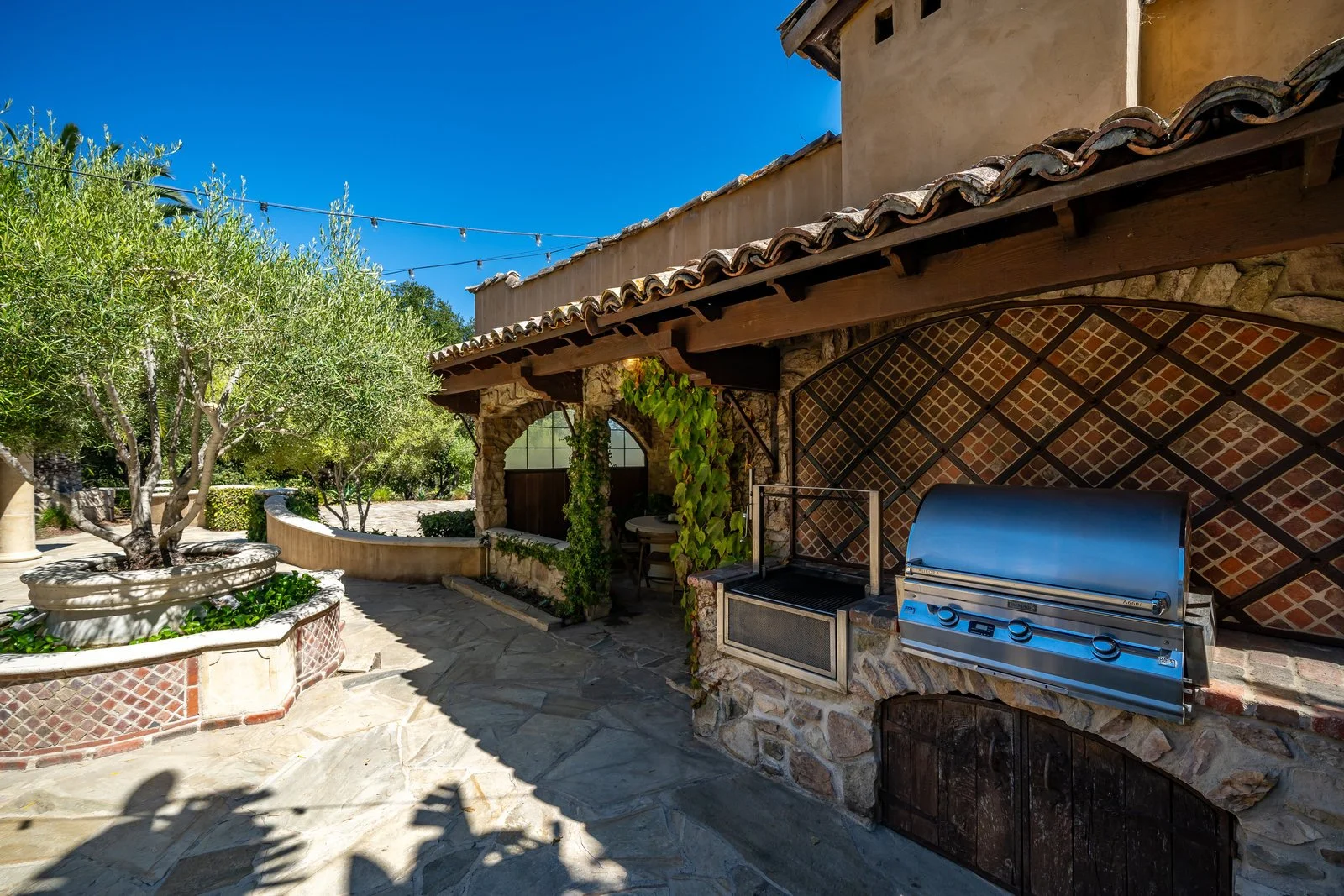 Outdoor patio area with trees, stone planters, a brick and stone fireplace, and a stainless steel grill under a tiled roof.