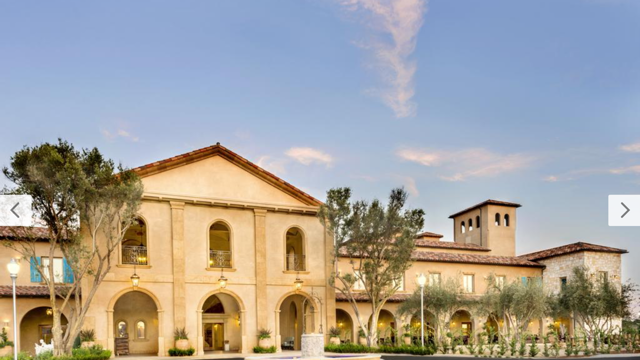 A large Mediterranean-style building with arched windows and doorways, surrounded by trees and a landscaped walkway, under a partly cloudy sky.