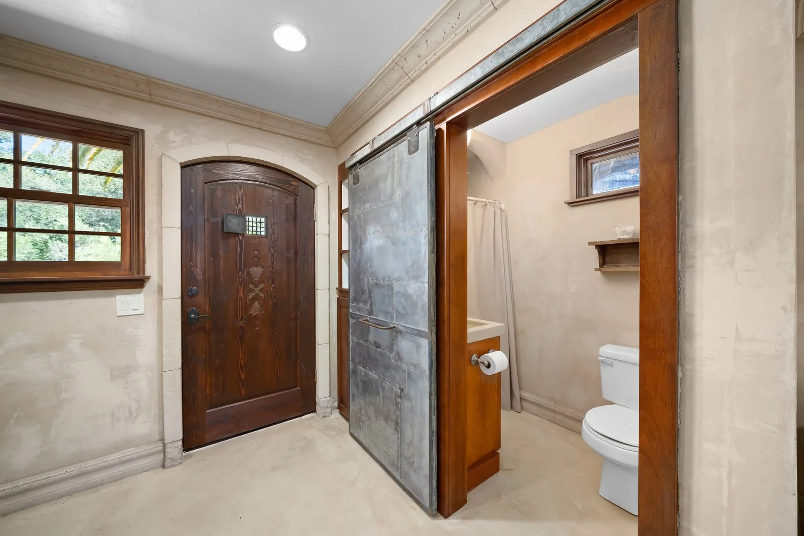 Bathroom entrance with wooden door, small window, and a bathroom with a toilet, shower, and wall shelf, in a rustic style.