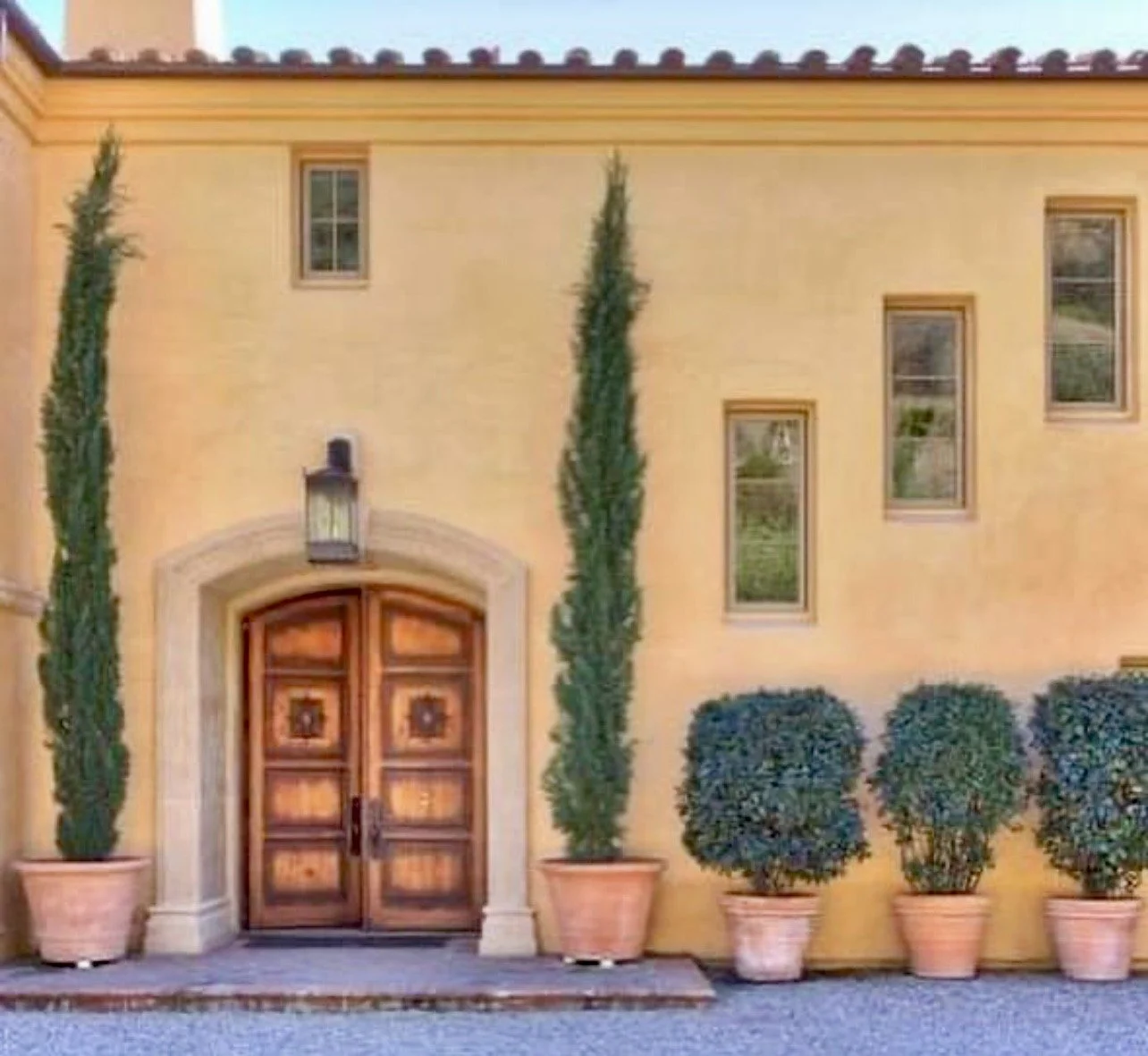 Front entrance of a yellow stucco house with a wooden double door, potted tall cypress trees on either side, and four rounded bushes in terracotta pots at the base, with three rectangular windows above.