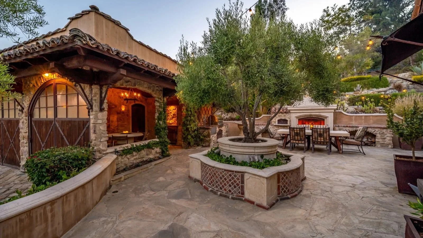 A cozy outdoor patio with a large central tree in a circular stone planter, surrounded by stone-paved flooring, outdoor seating, and a built-in fireplace with a glowing fire. There are string lights overhead and greenery around the space.