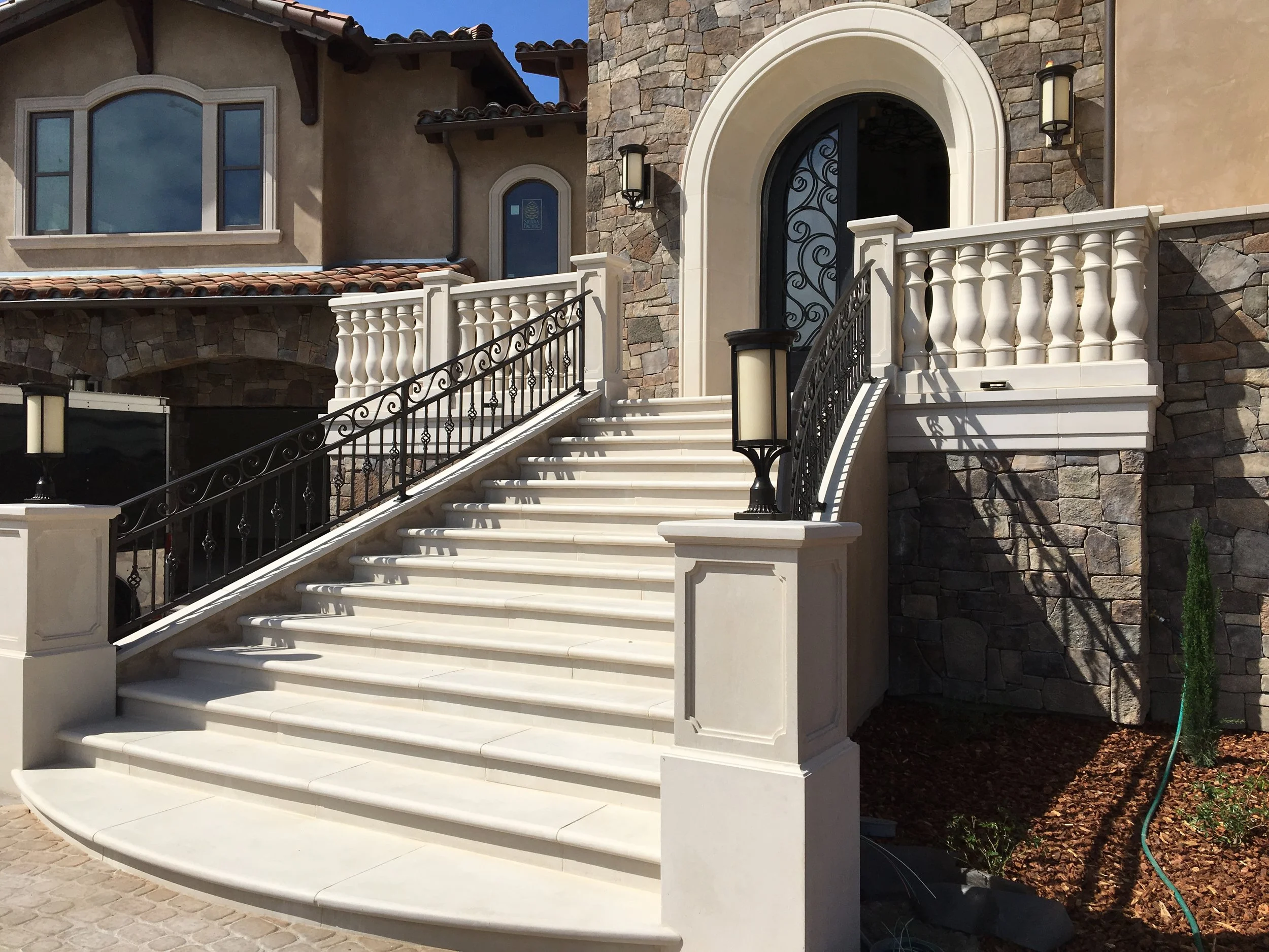 Front entrance of a luxurious house with white stairs, black wrought iron railings, and a stone wall, featuring two wall-mounted lanterns and an arched door.