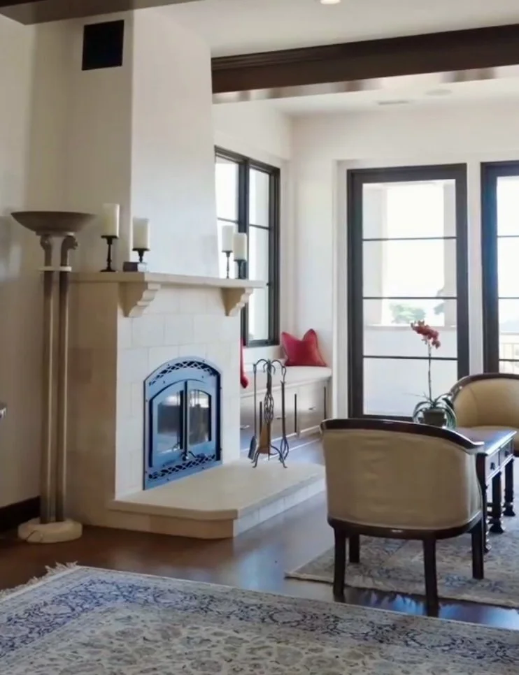 Living room with a fireplace, large windows, a built-in window seat with red pillows, a potted orchid, chairs, a fireplace tool set, candles on the mantel, and a carpeted floor.