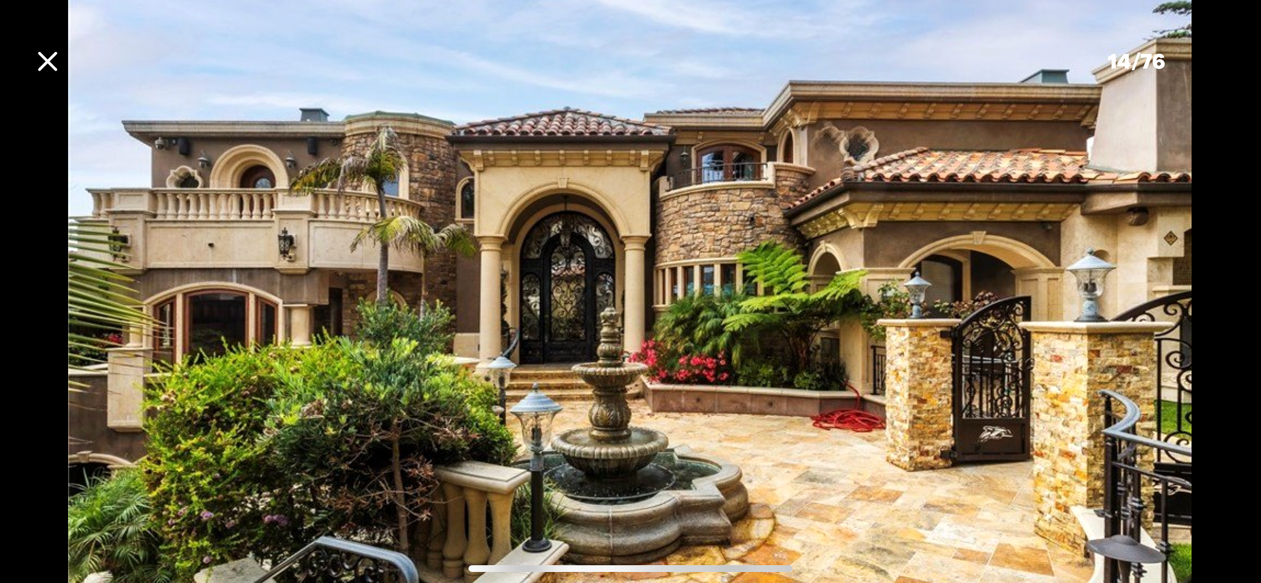 Luxurious multi-story house with stone and stucco exterior, tiled roof, decorative iron gates, lush landscaping, fountain, and outdoor lighting.