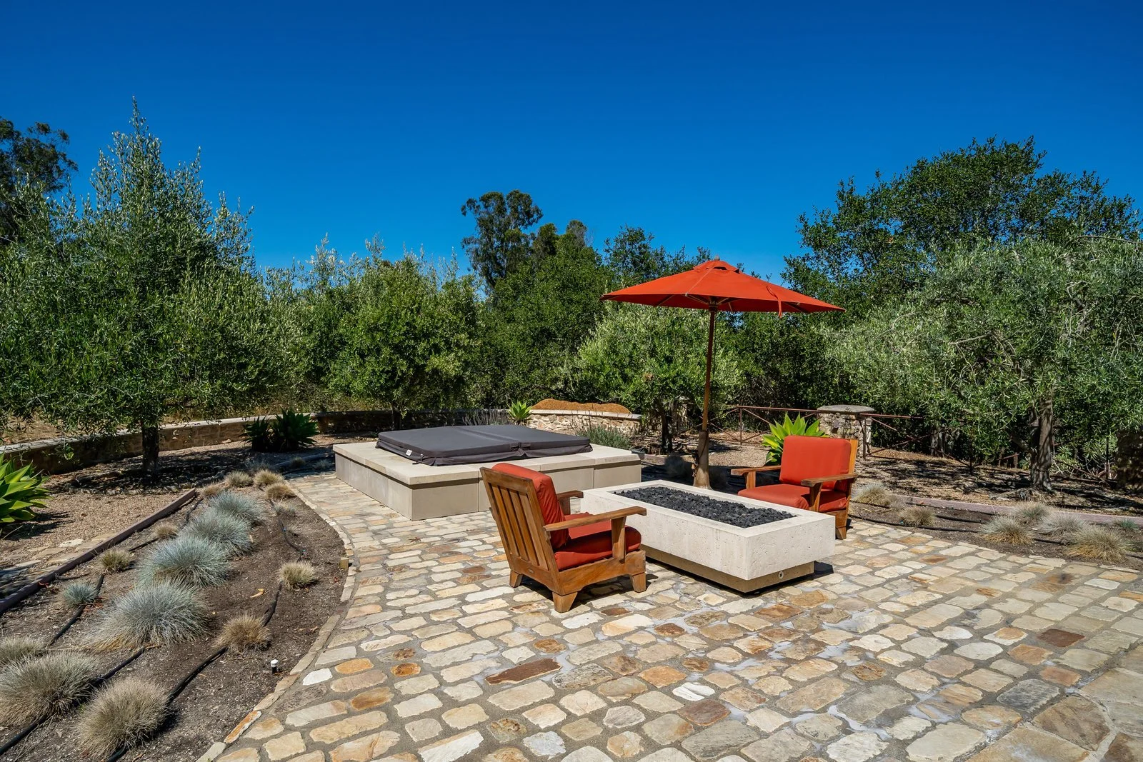 Outdoor patio with stone pavers, orange umbrella, wooden chairs with orange cushions, a fire pit, and a hot tub surrounded by greenery and trees under a clear blue sky.