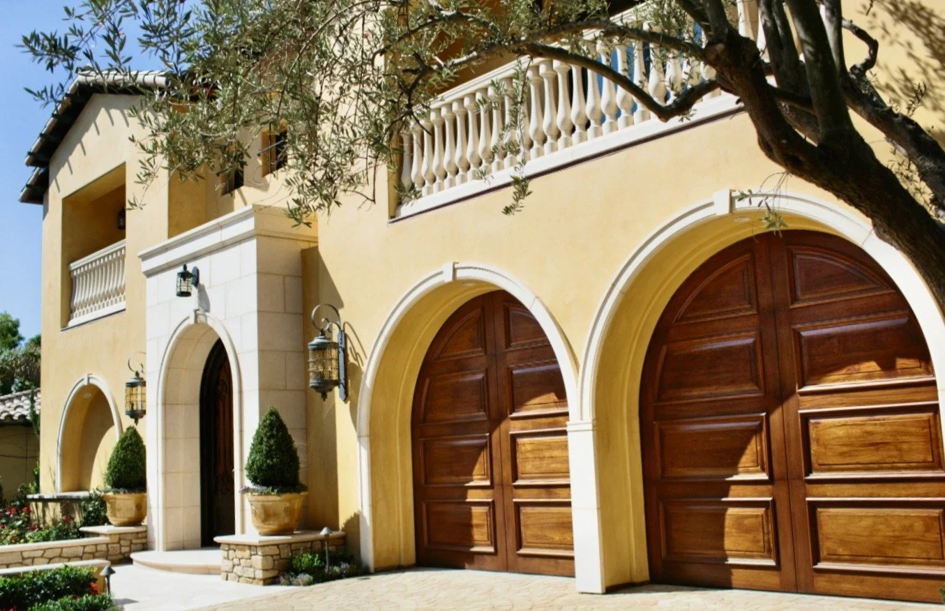 Luxury house with arched wooden garage doors, balconies, and potted plants, surrounded by trees.
