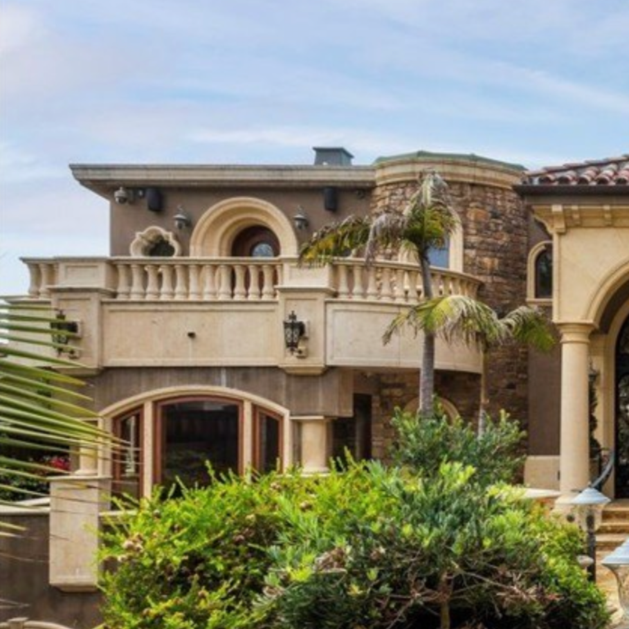 Exterior of a luxury house with a balcony, arched windows, and decorative architecture, surrounded by palm trees and greenery.
