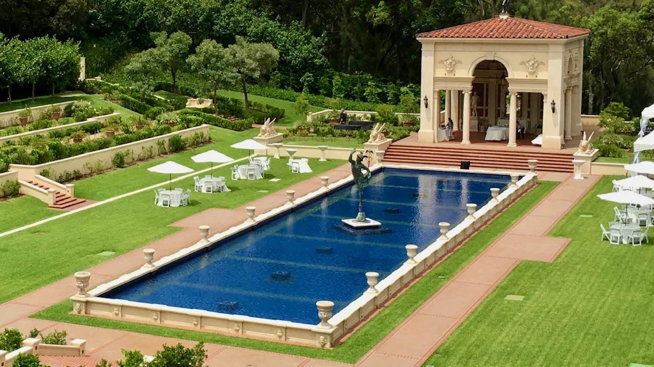 A large outdoor area with a rectangular blue pond, a small classical pavilion with columns, statue fountain in the middle of the pond, surrounded by green lawn, white chairs and umbrellas, and lush trees in the background.