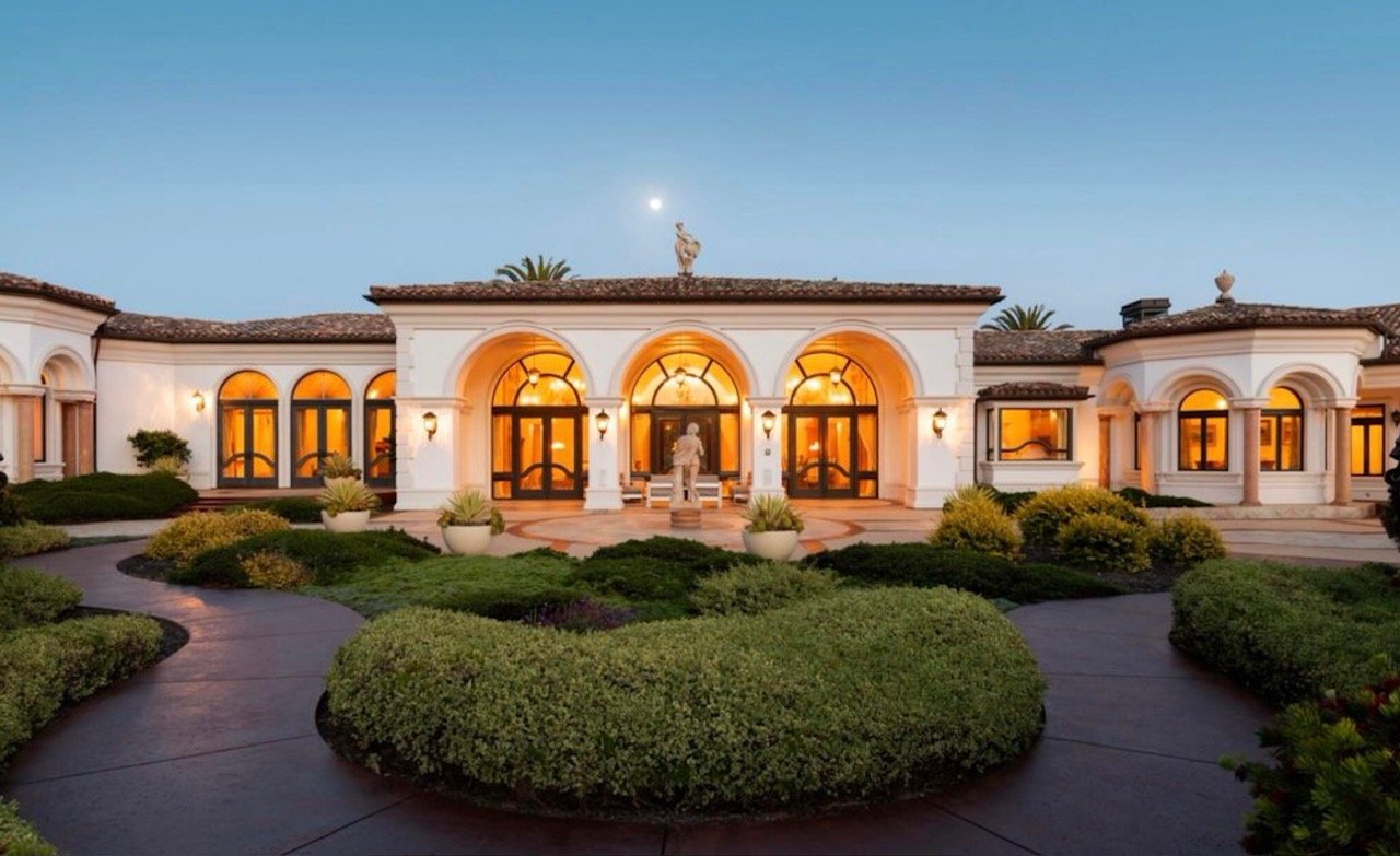 Luxury mansion with arched windows and a manicured garden in the evening, moon visible in the sky.