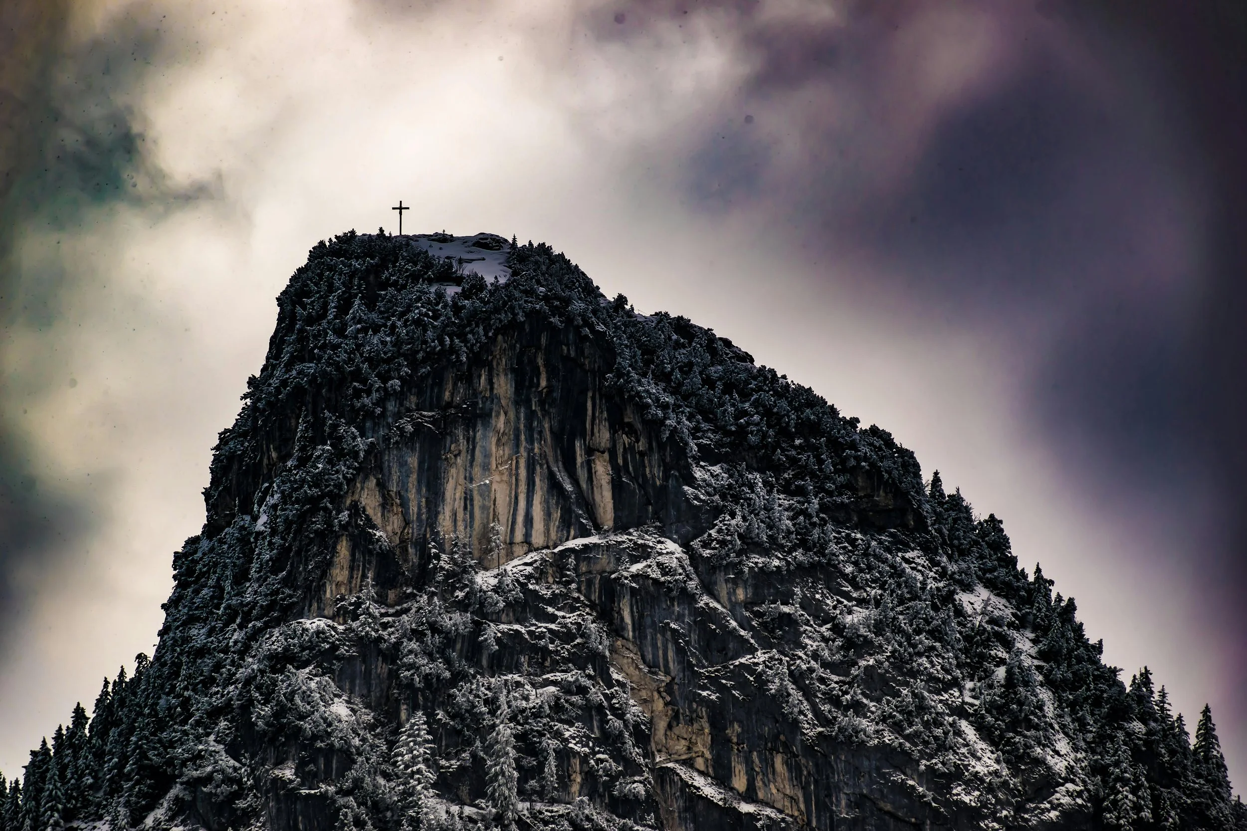A snow-covered mountain peak with a cross at the summit and a cloudy sky in the background.