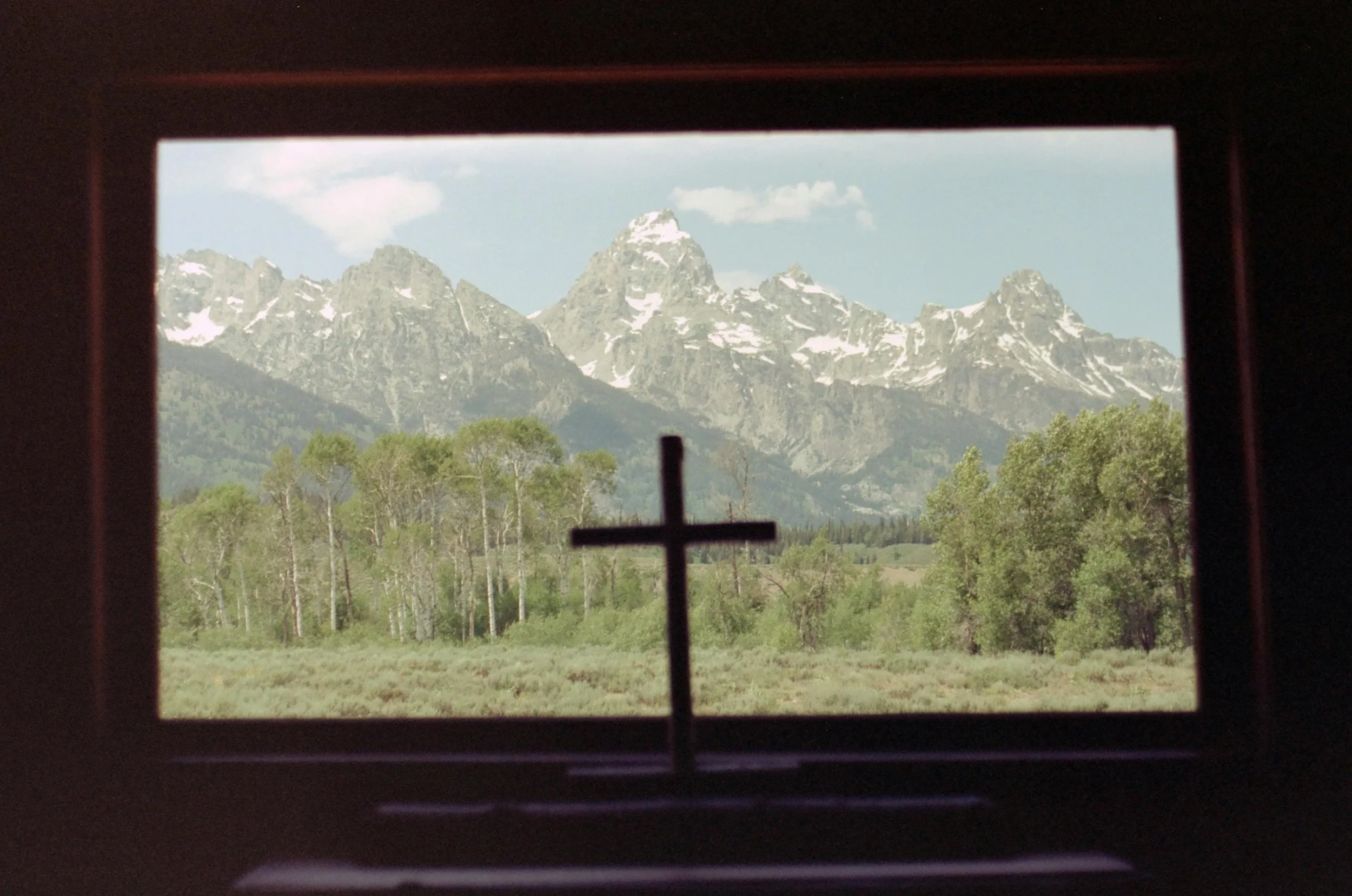 View through a window with a small cross-shaped religious symbol in the foreground, showing a landscape of green trees and snow-capped mountains in the background.
