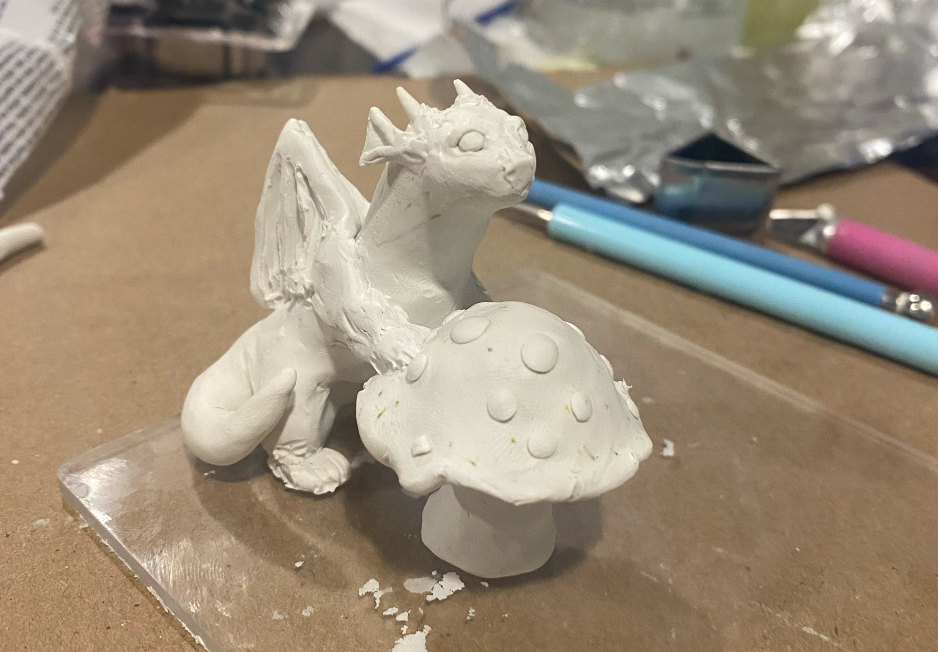 Unfinished polymer clay dragon sculpture of a tiny dragon leaning against a spotted mushroom.