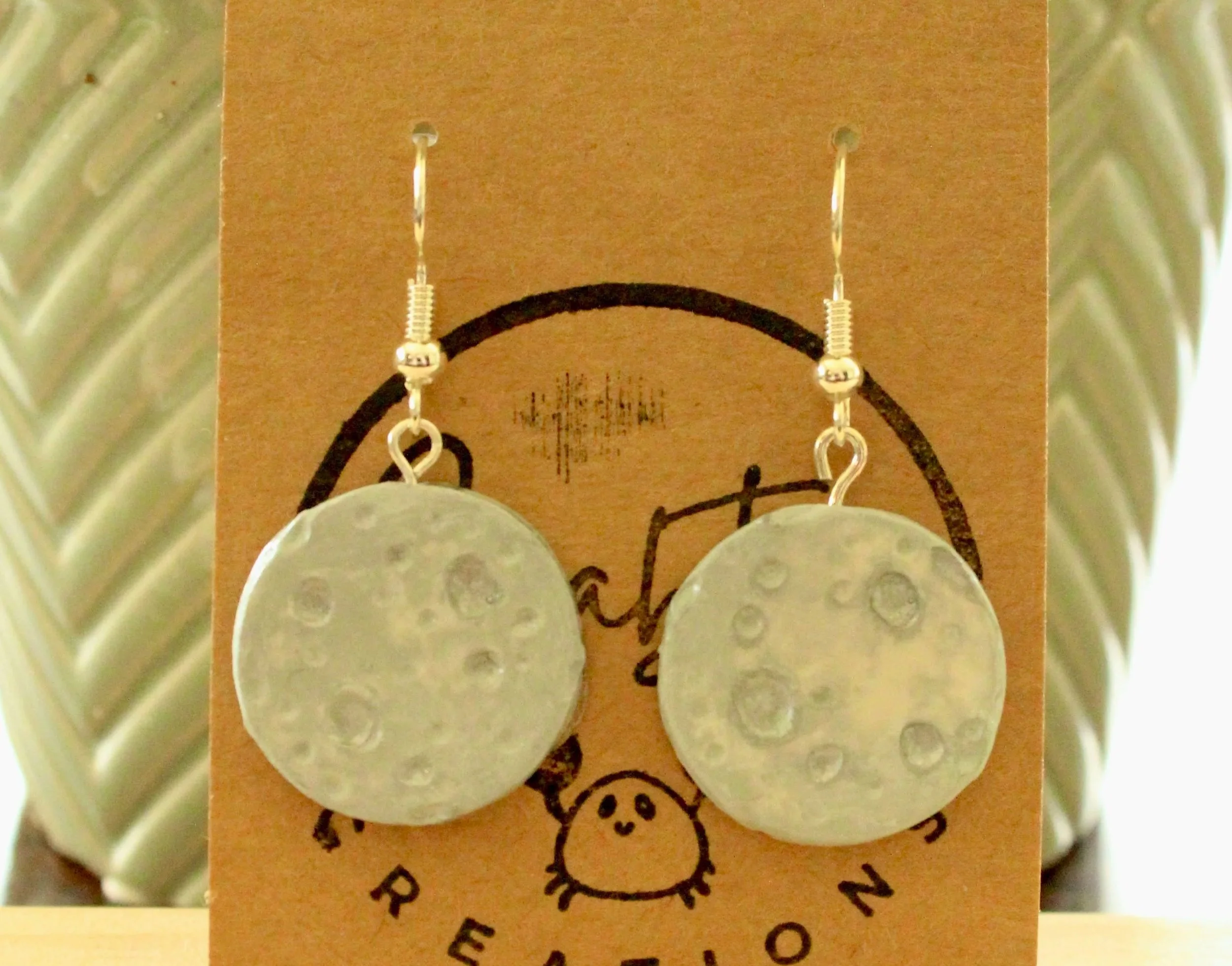 Handmade polymer clay moon earrings, dangly earrings, against a cardboard backing and leaned against a potted houseplant.