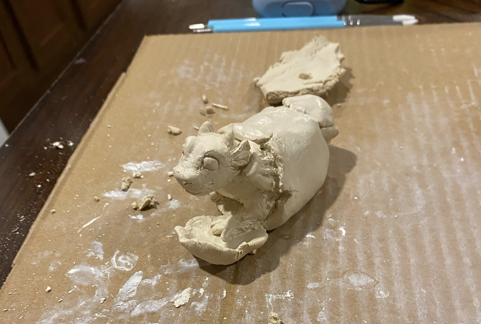 Unfinished dragon sculpture of a baby dragon hatching out of an egg, sitting atop a work surface and surrounded by unused pieces of clay and sculpting tools.