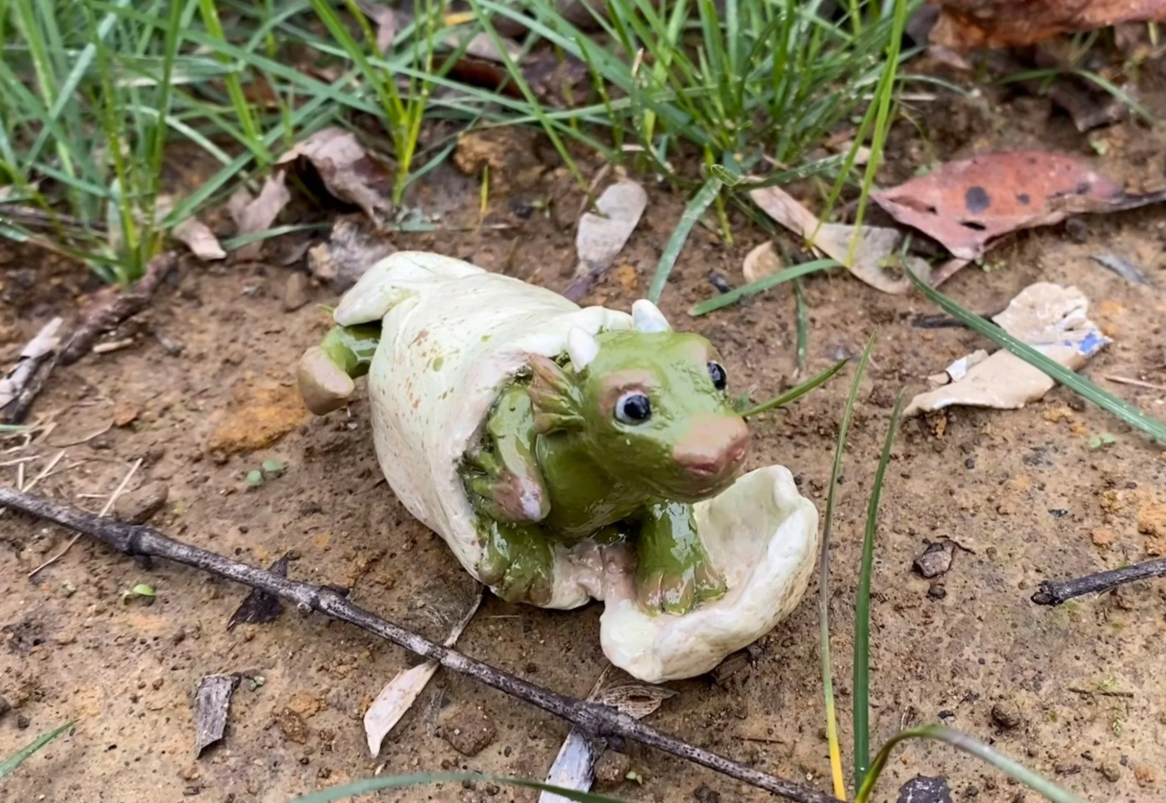 Polymer clay sculpture of a baby dragon hatching from its shell, sitting on the ground outside surrounded by grass, sticks, and leaves.