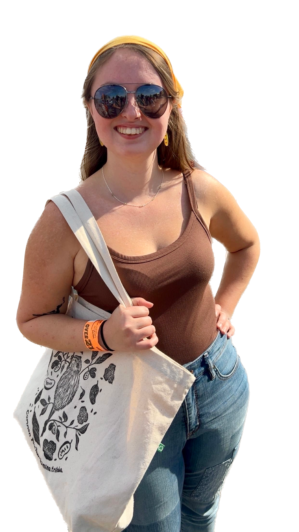 Adrianne Crabtree, artist of Crabtree Creations. A woman smiling outdoors wearing sunglasses, a yellow headband, a brown sleeveless top, blue jeans, and carrying a canvas tote bag with a bird and floral design.