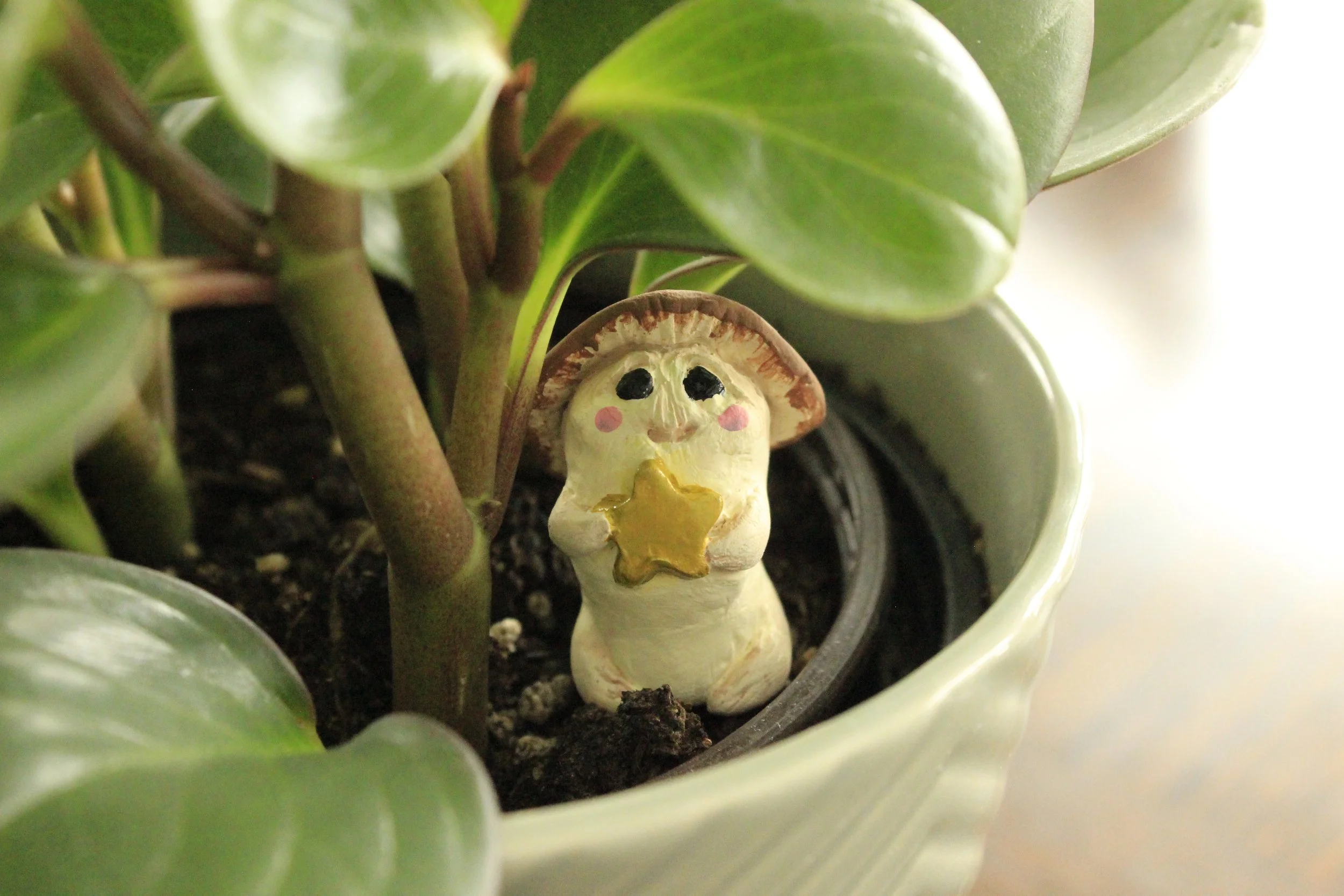 Polymer clay sculpture of a mushroom folk, or mushroom man, holding a gold star while sitting beneath the leaves of a potted house plant.