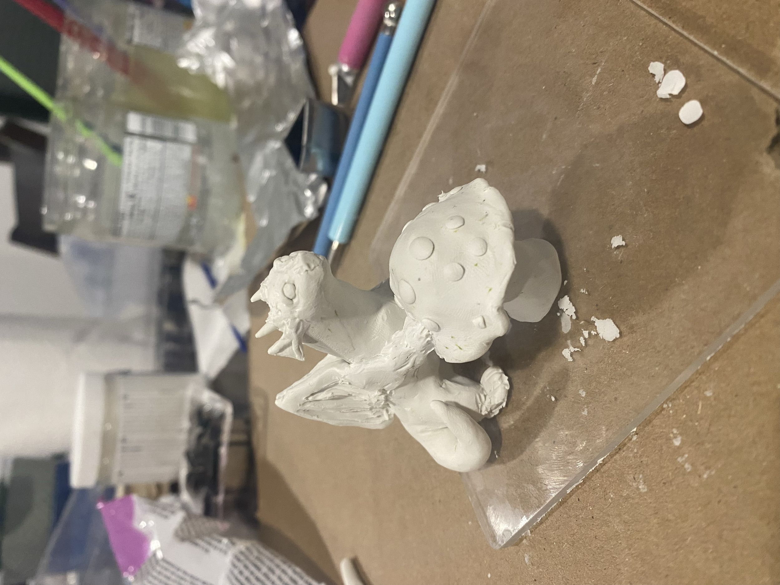 Unfinished polymer clay statue of a tiny dragon leaning against a mushroom, this sculpture is sitting atop a workstation with sculpting tools and paintbrushes.