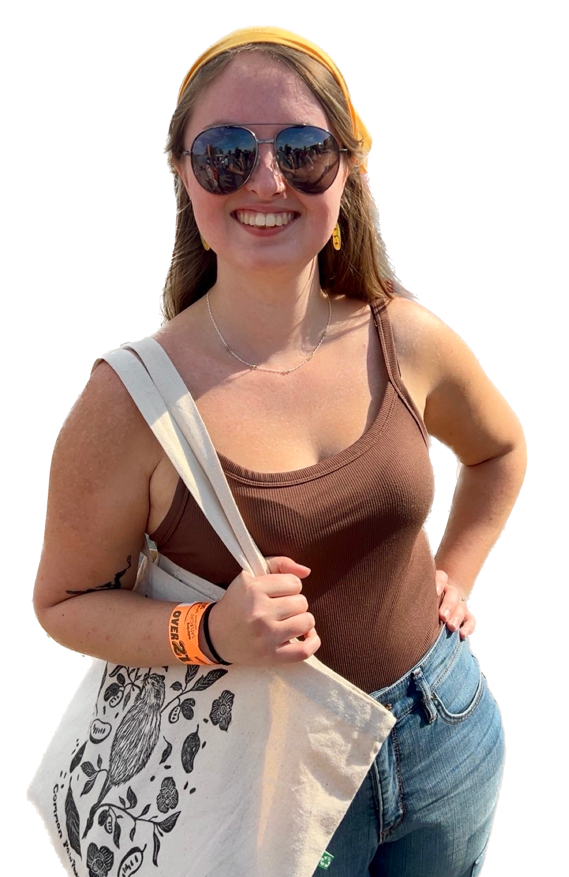Adrianne Crabtree, artist of Crabtree Creations. A woman smiling outdoors wearing sunglasses, a yellow headband, a brown sleeveless top, blue jeans, and carrying a canvas tote bag with a bird and floral design.