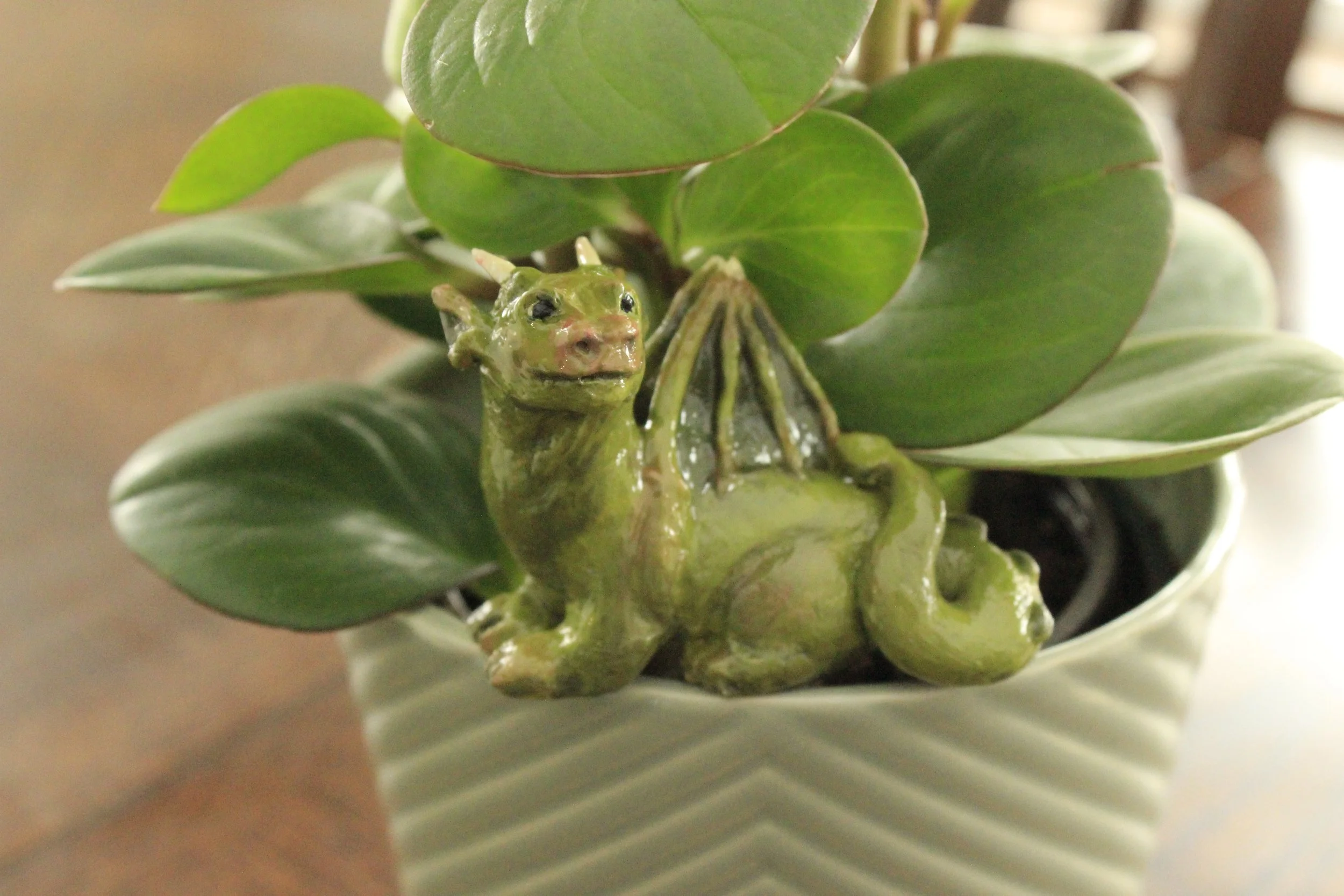 A polymer clay dragon sculpture sitting amongst the leaves of a potted houseplant atop a wooden table surface.