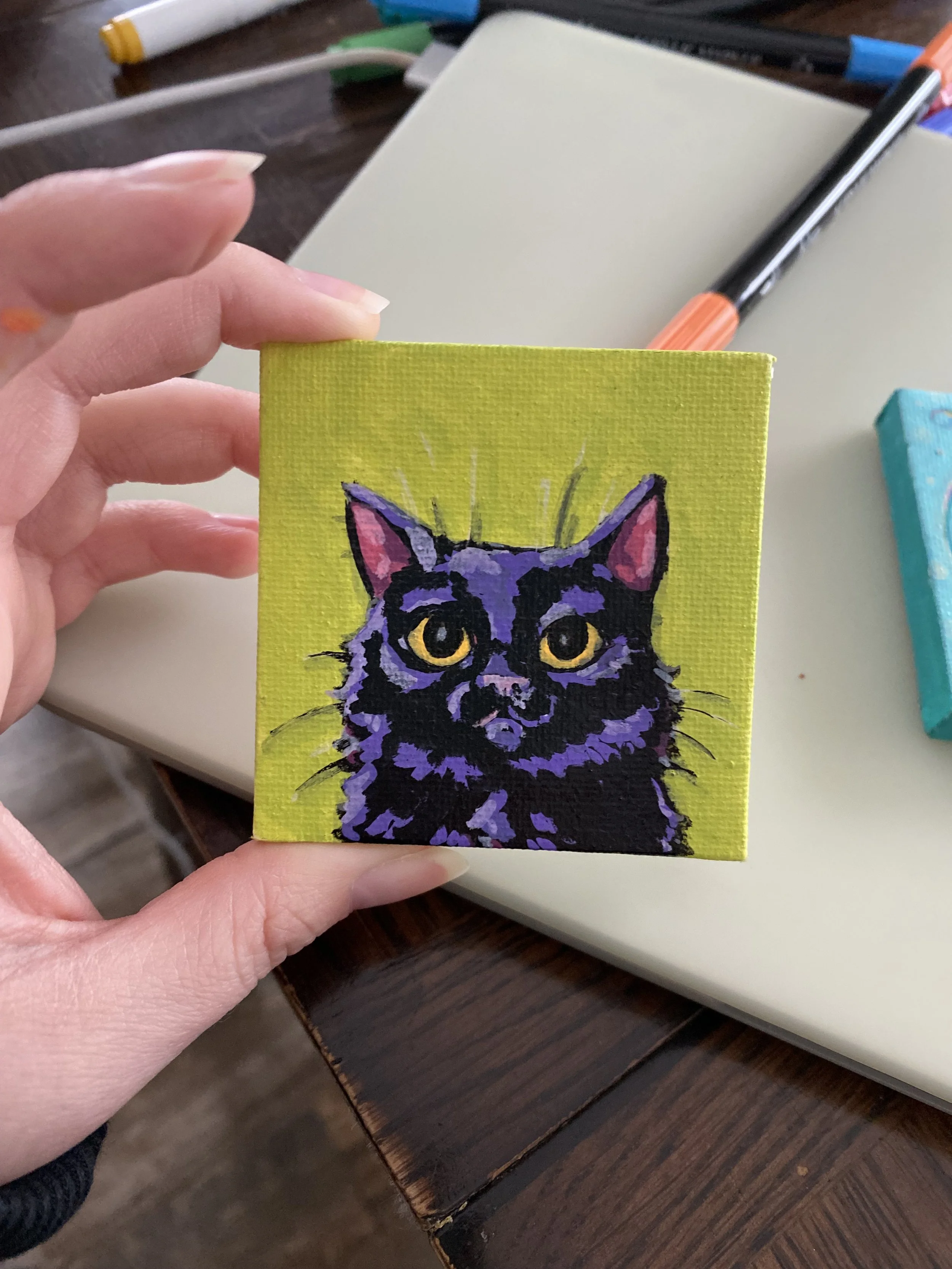 A miniature hand-painted canvas of a black and purple cat with yellow eyes being held in the hand of the female artist.