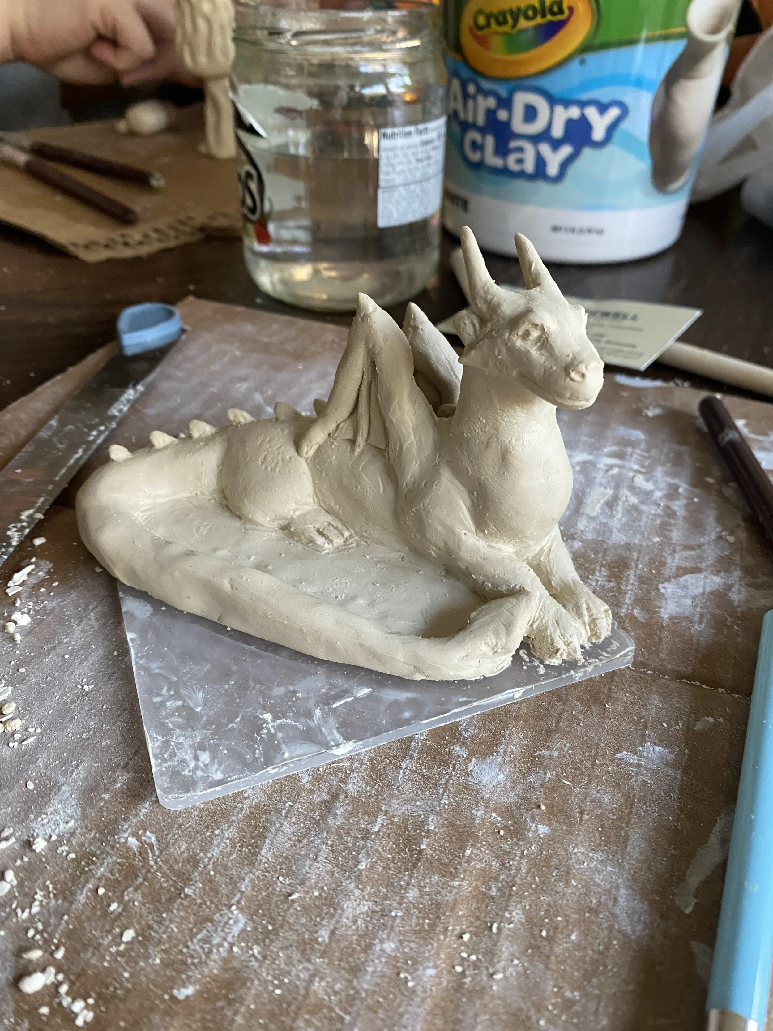 Unfinished sculpture of a dragon business card holder sitting atop a workspace and surrounded buy sculpting tools, clay, and clay cutters.