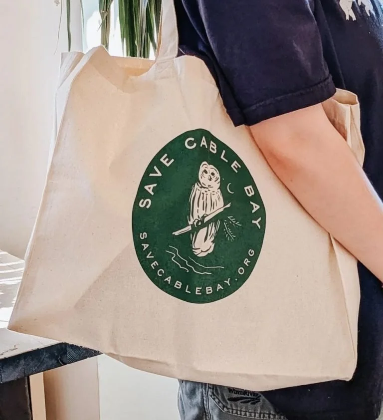 Person holding a beige reusable shopping bag with a green circular logo that features an otter holding a stick, with the text 'Save Cable Bay' and 'savecablebay.org' around the logo.