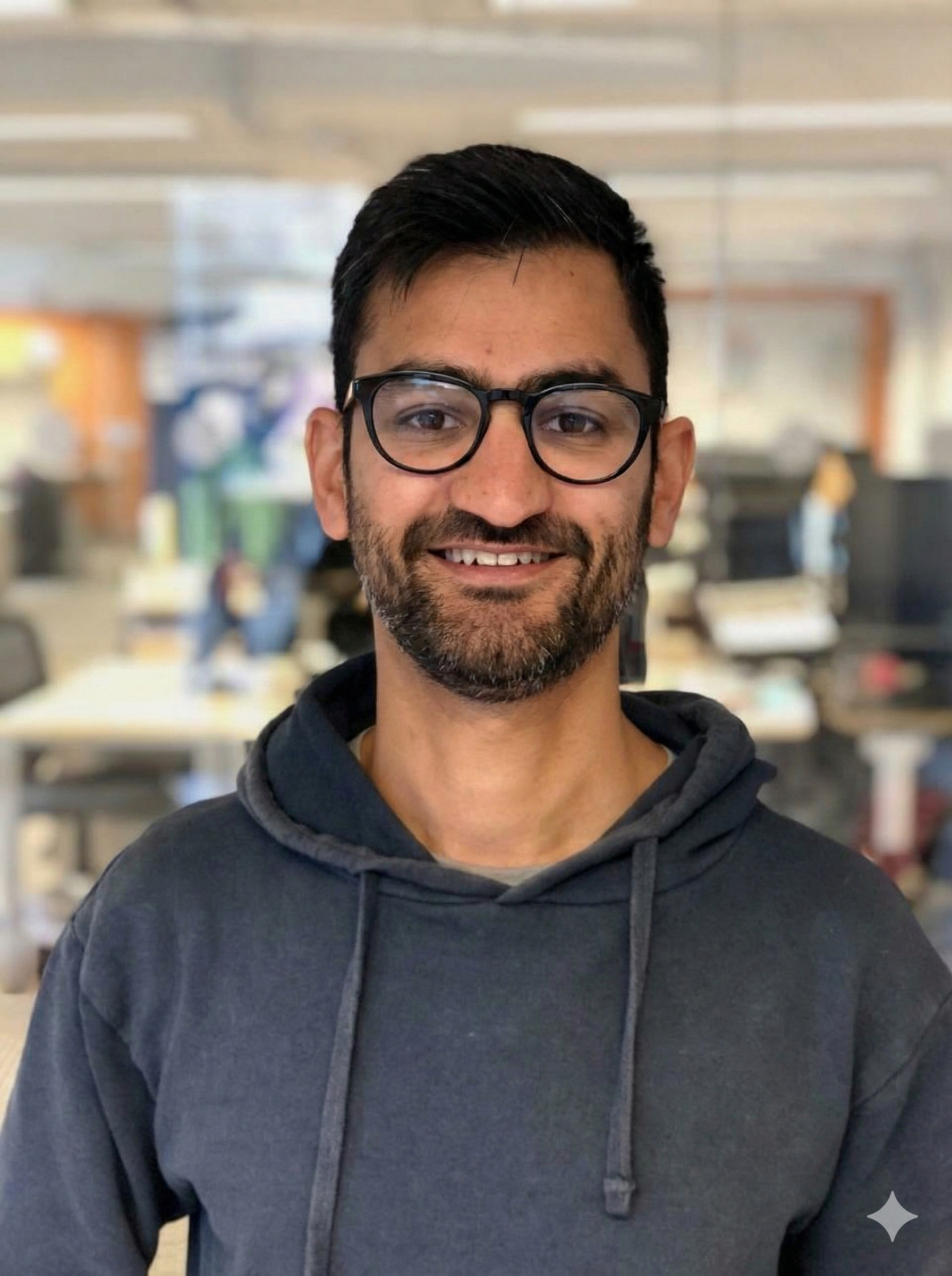 A man with short dark hair, glasses, and a beard, smiling in an office setting, wearing a dark hoodie.