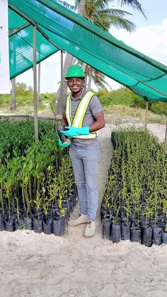 A man smiling in a green safety helmet and reflective vest, standing between rows of young plants in nursery bags, holding a device, under a green shade cloth in a tropical outdoor setting.