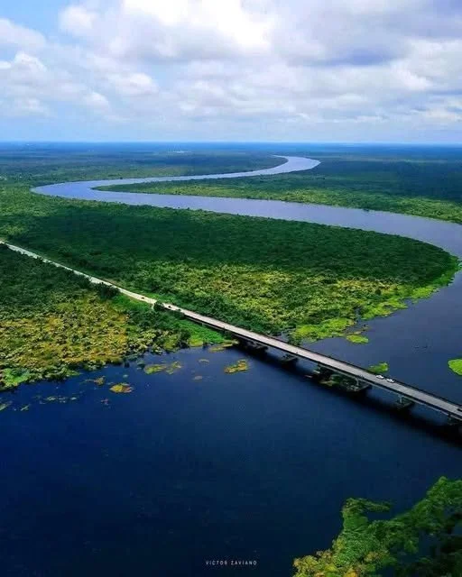 Aerial view of a winding river flowing through a green landscape, with a bridge crossing over the water and a road running parallel to it.
