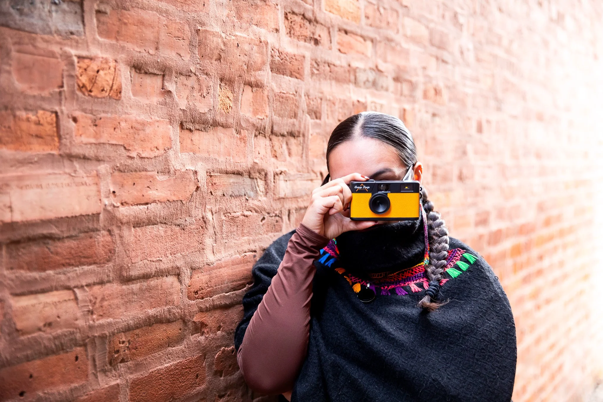 Angela Estrella Mejia, founder of Angie Star Studios, holding a yellow camera and photographing in a brick alleyway, representing her creative practice in photography and visual storytelling.