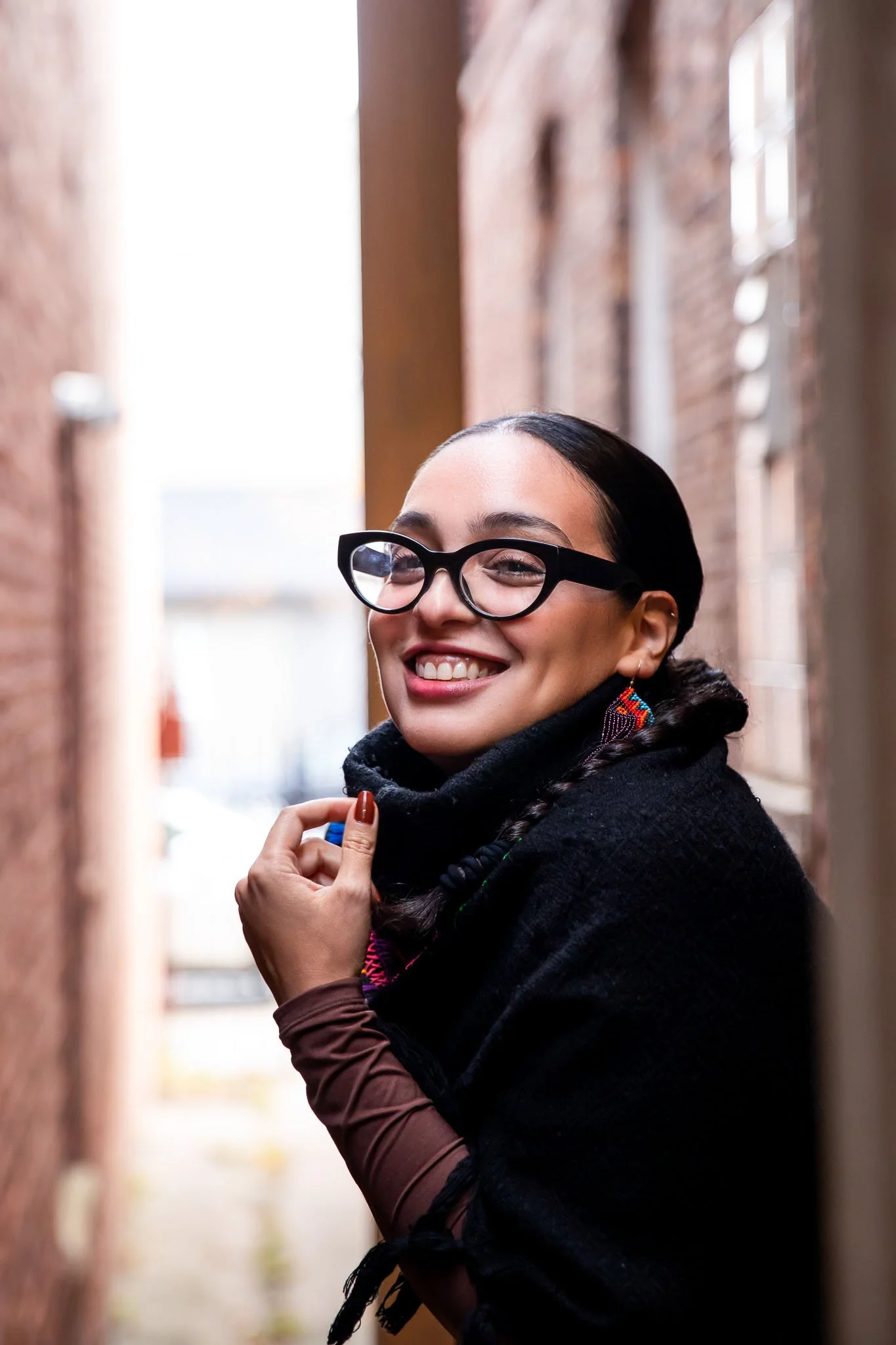 Portrait of Angela Estrella Mejia, founder of Angie Star Studios, smiling in an urban alleyway, wearing black glasses and a shawl, reflecting a warm, creative, and approachable presence.