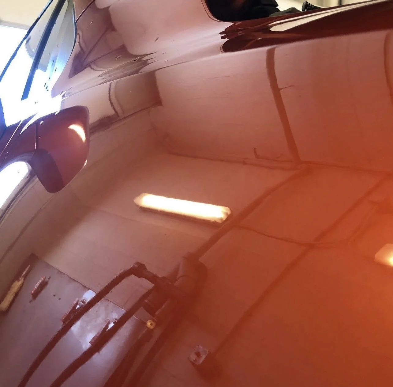 Close-up view of a shiny, reddish-brown car's body with reflections of the garage interior and fluorescent lights.