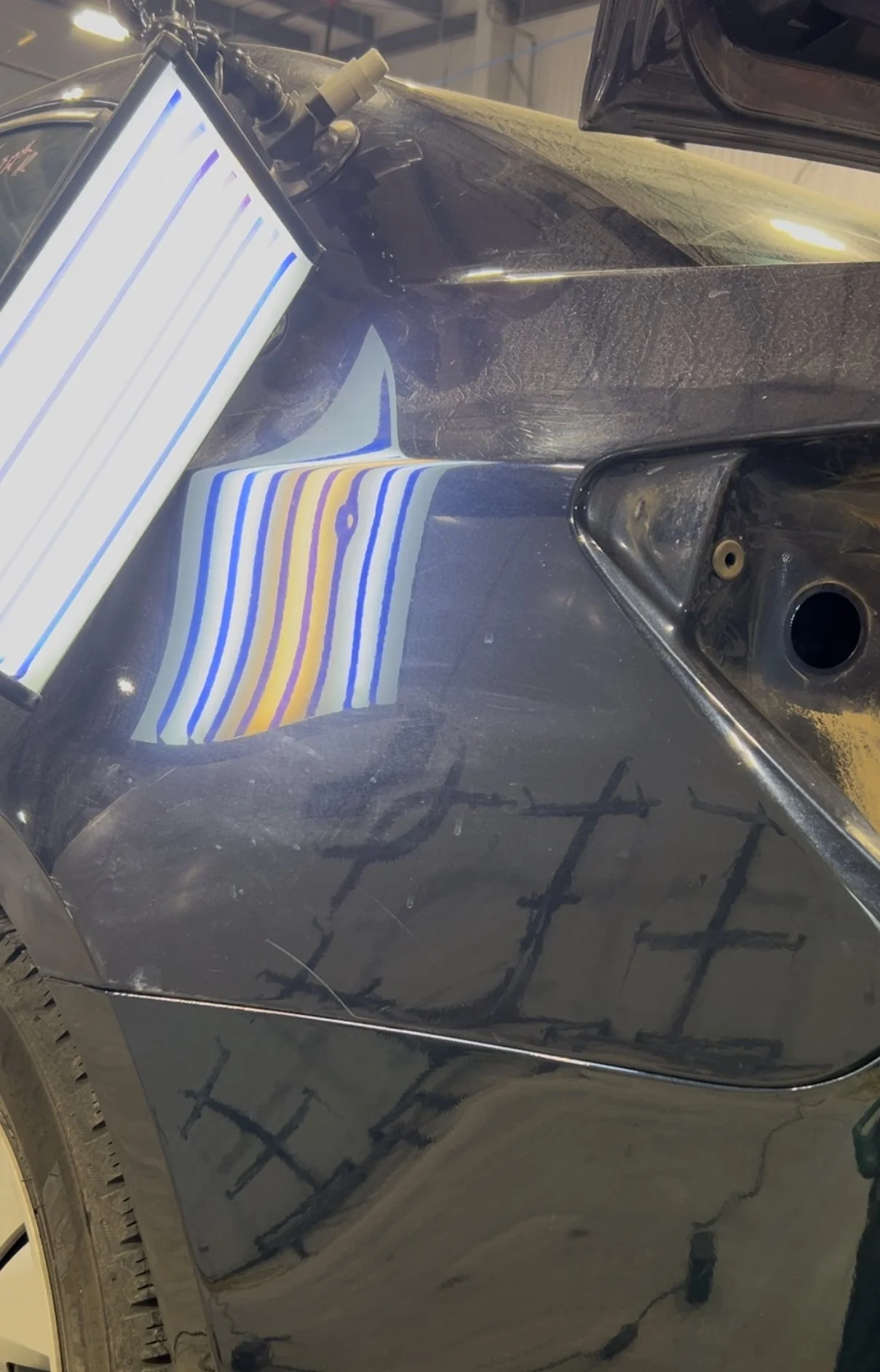 Close-up of a black car's dented and scratched side with some markings, a striped sticker, and a digital device attached to the car's surface.