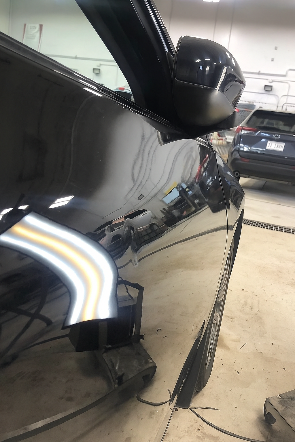 Close-up of a black car's side mirror and door, with reflections of other vehicles and the indoor garage.